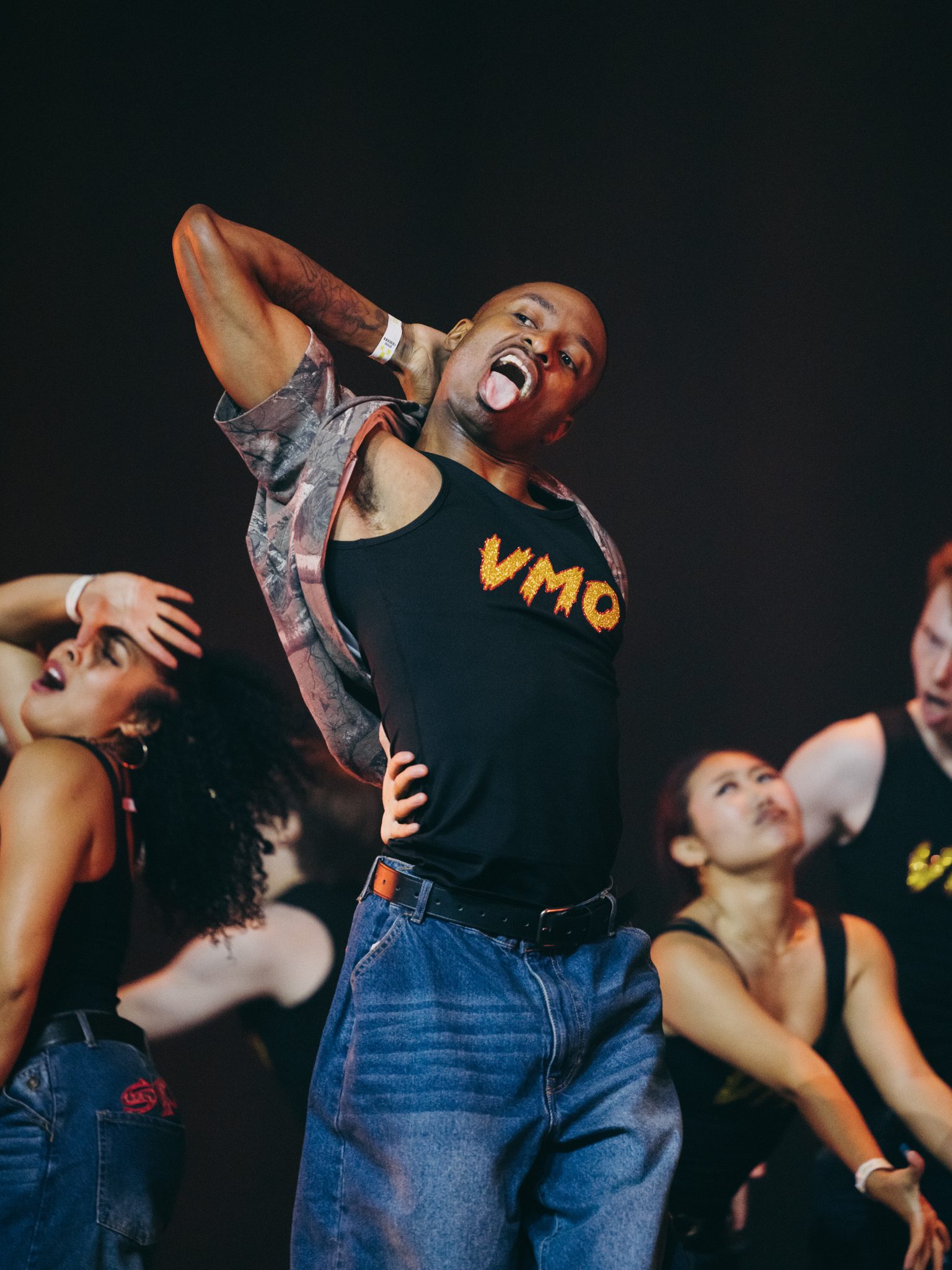 Rapper performing on stage in black vest with yellow "VMQ" text, surrounded by dancers against dark background.