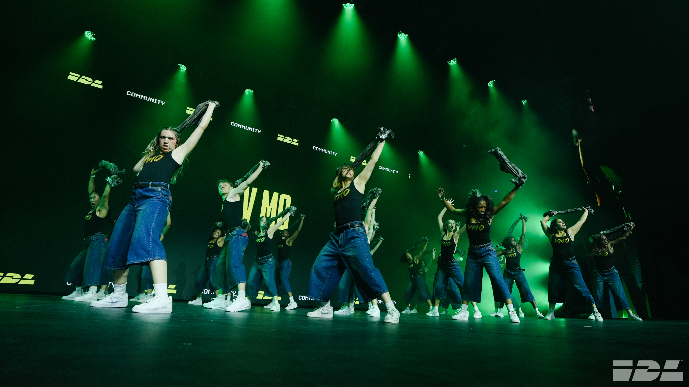 Dance group in blue outfits performing on stage with bright green stage lighting and smoke effects in dark venue.