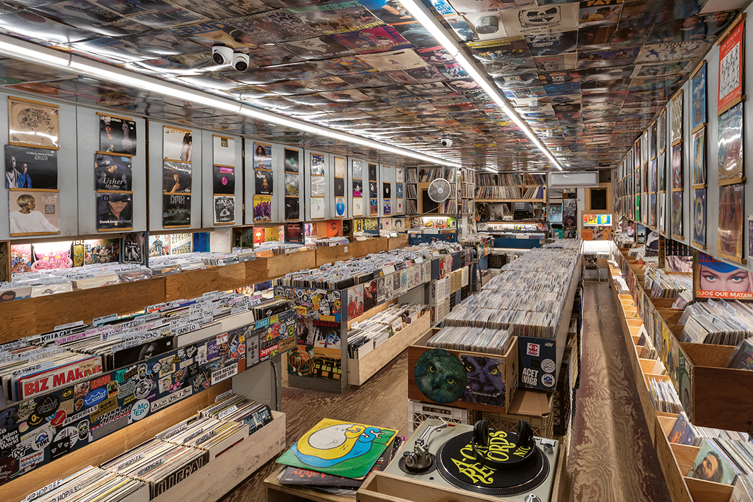 Record shop interior with wooden bins full of vinyl records, fluorescent lighting, and walls covered in album artwork and posters.