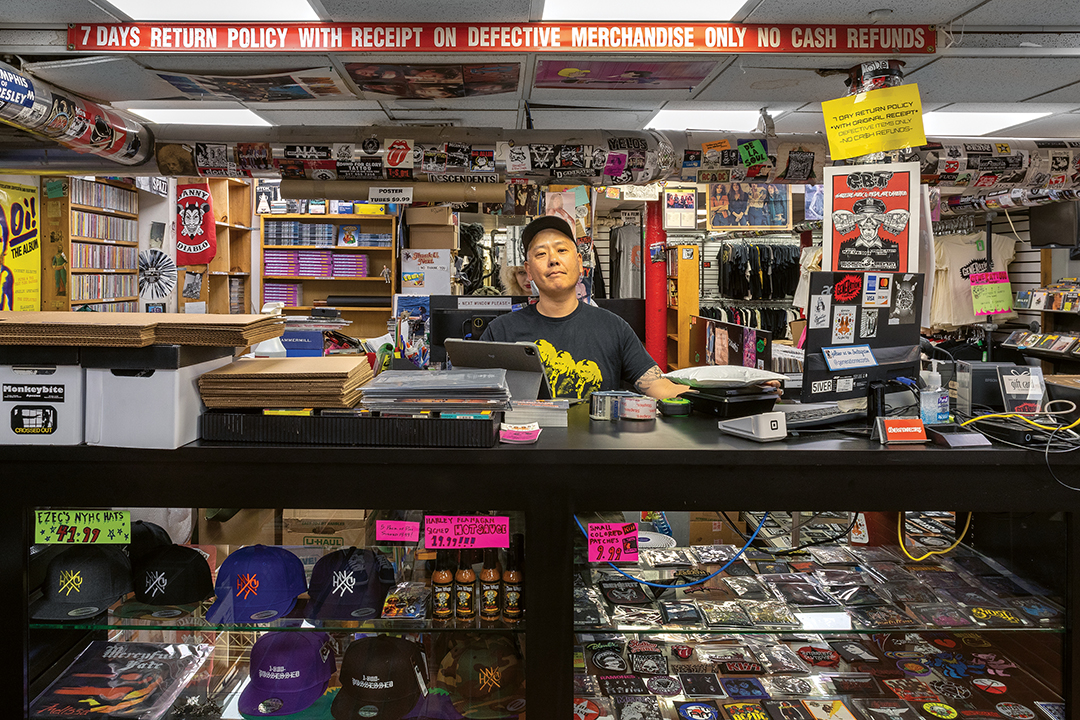 Man behind cluttered shop counter with merchandise displays, shelving units, and red return policy banner overhead.