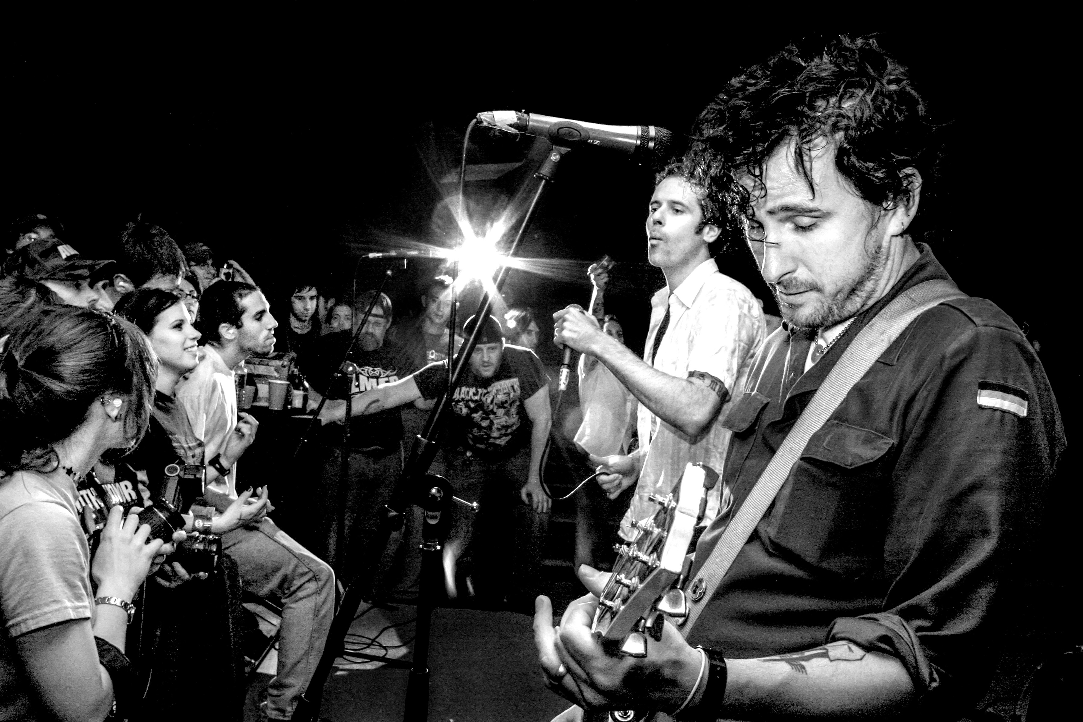 Black and white image of musicians performing in small venue. Guitarist with curly hair in foreground, singer at microphone behind. Seated audience watching from left side.