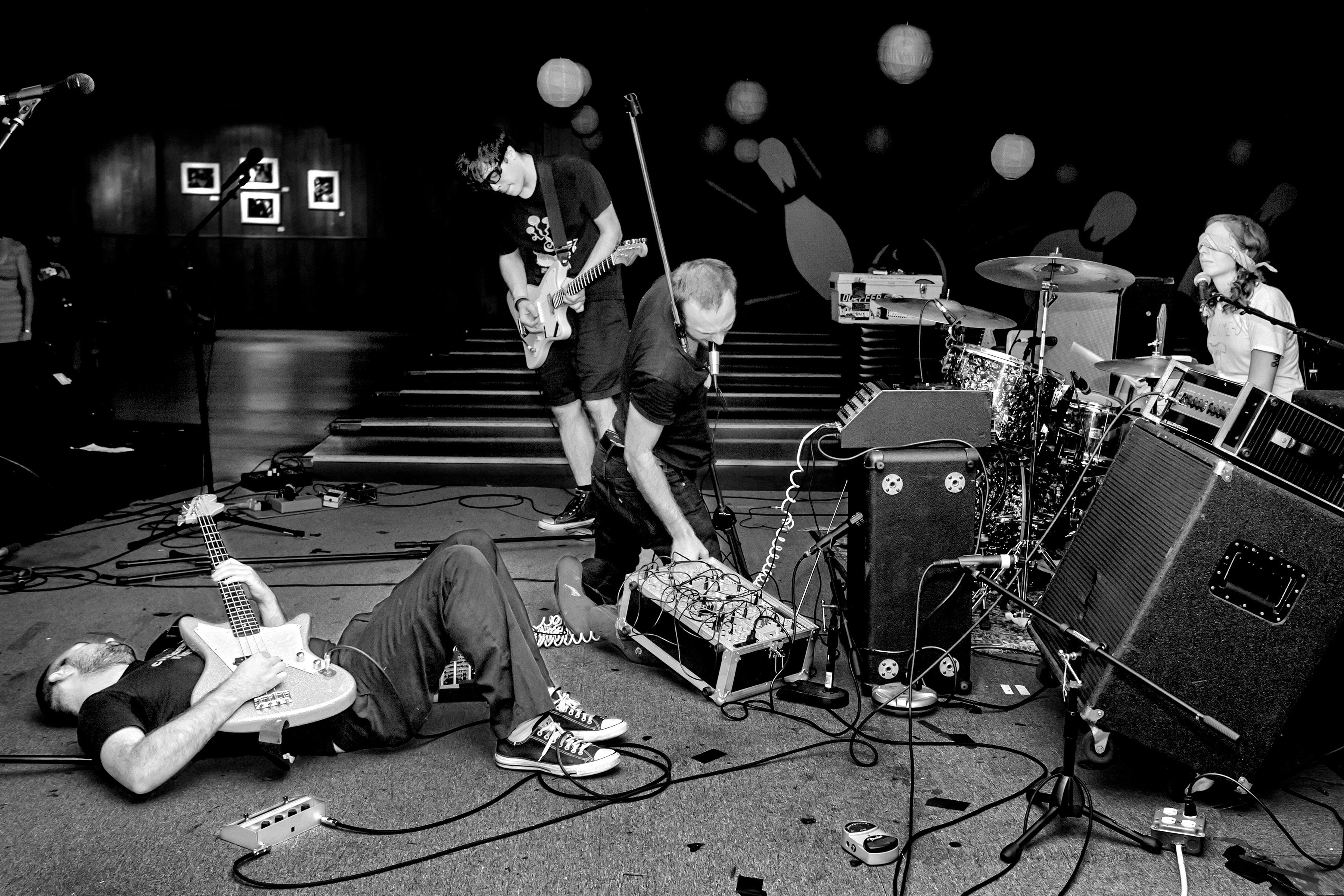 Black and white concert scene with band members on stage, one lying on floor, amplifiers, drum kit, and stage lighting visible.