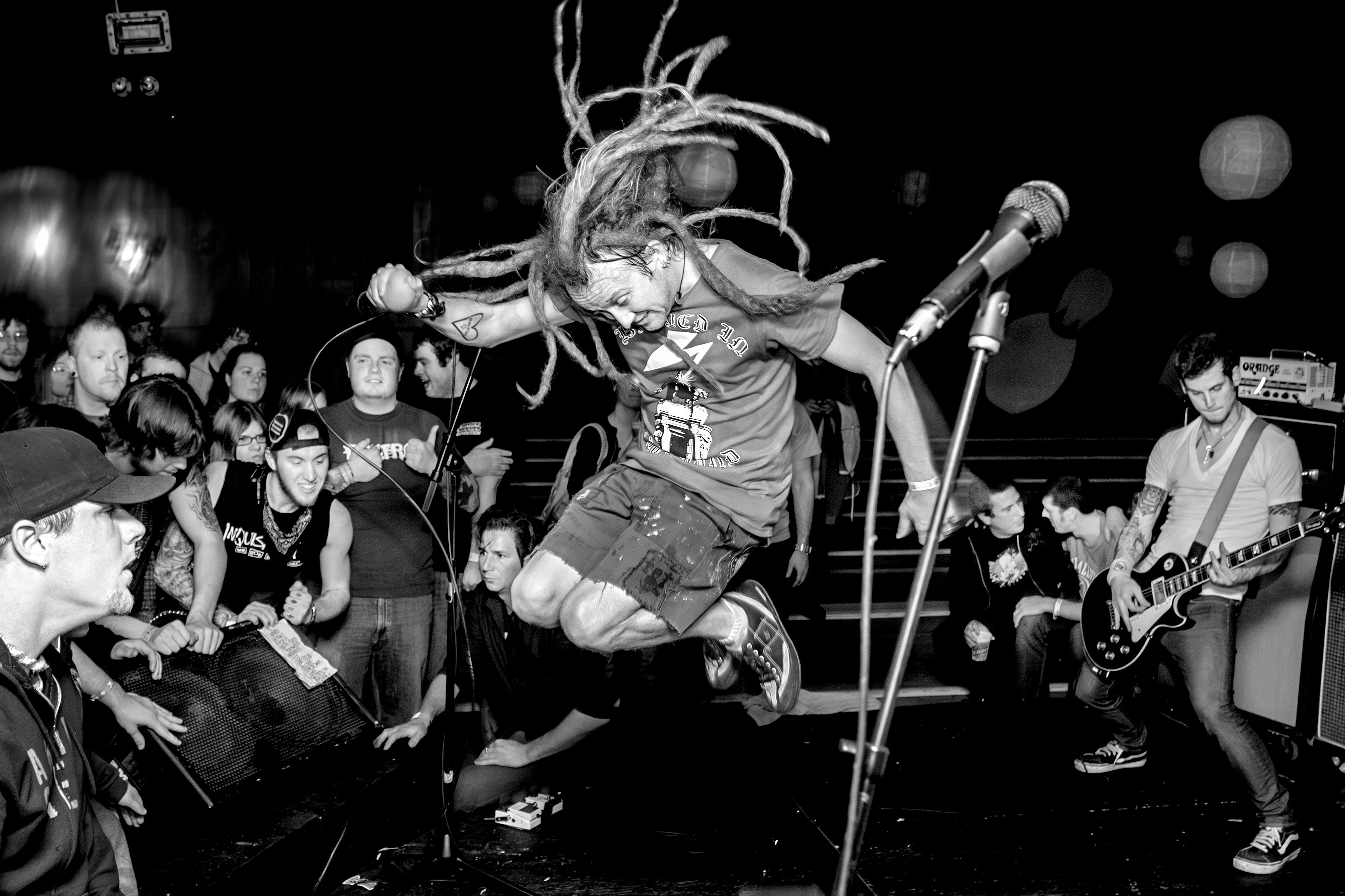 Black and white image of performer with dreadlocks jumping mid-air at microphone stand, surrounded by seated and standing audience members.