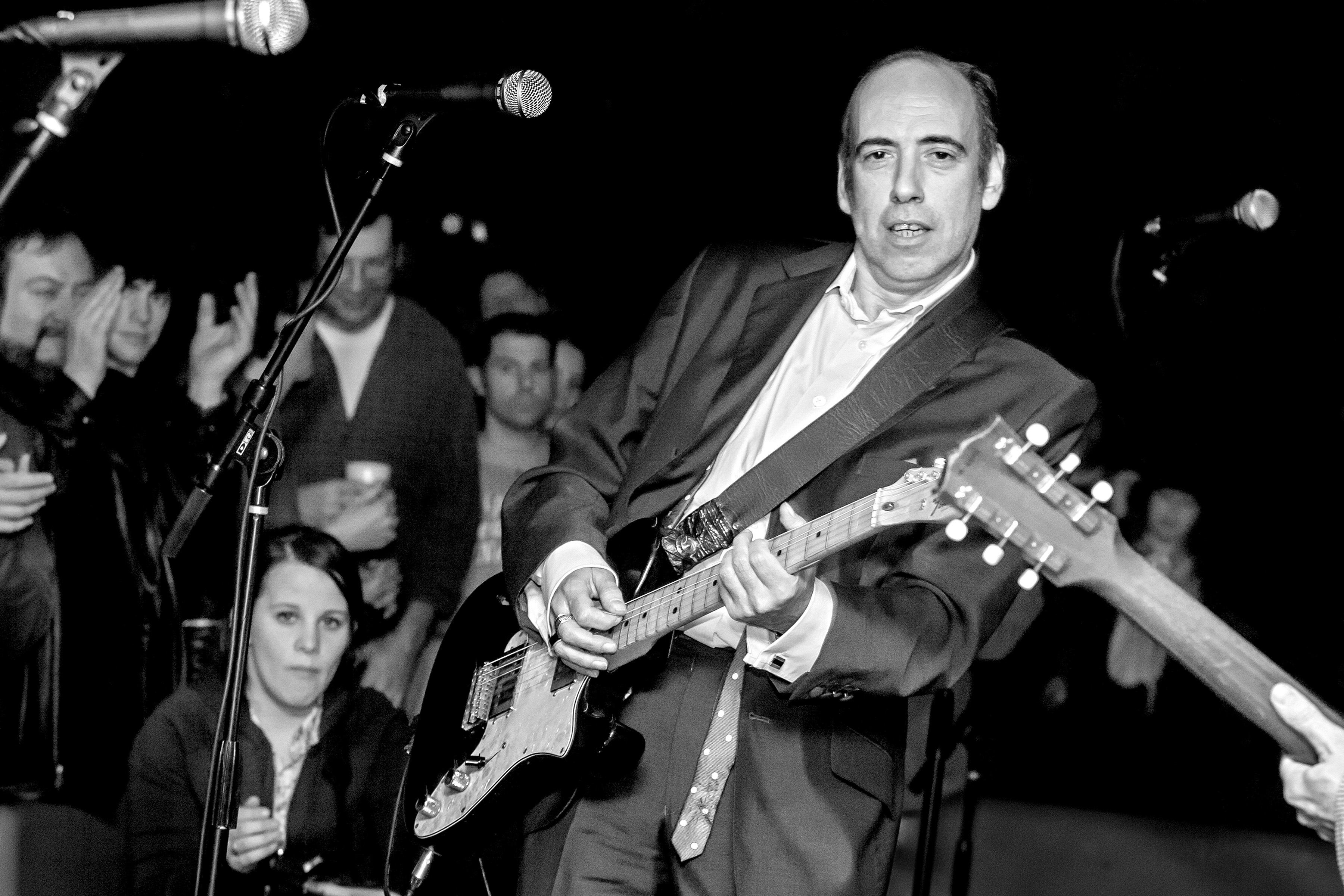 Black and white image of man in suit playing electric guitar on stage with microphones and audience in background.