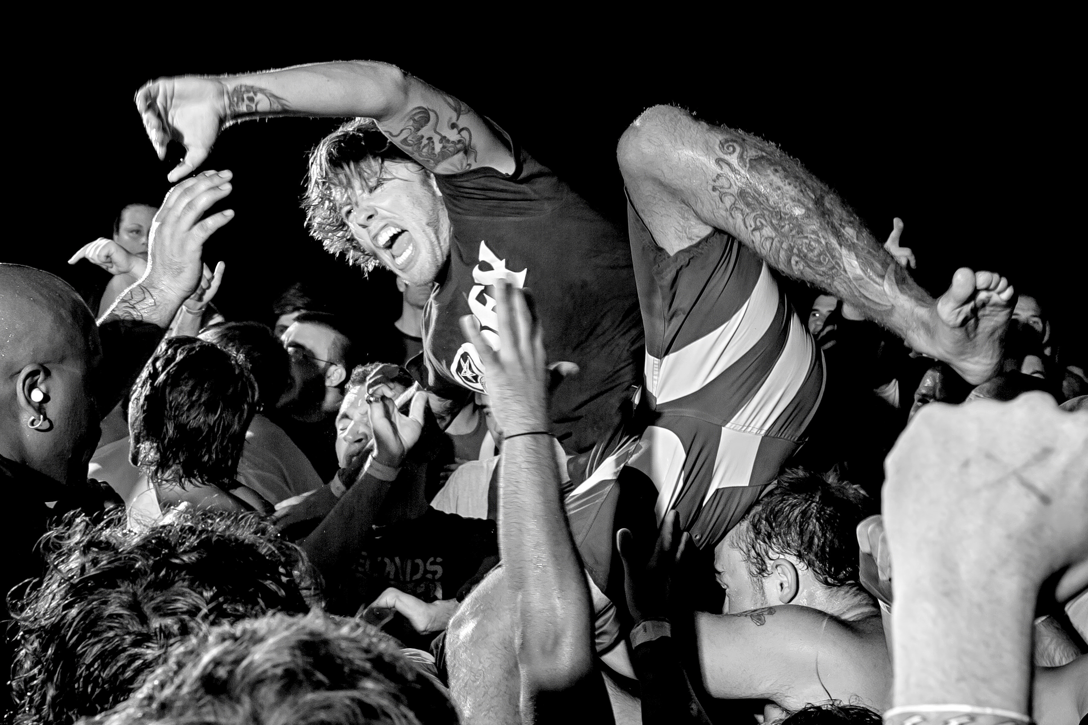 Black and white image of crowd surfing at concert, person in striped shirt carried overhead by multiple hands and arms.