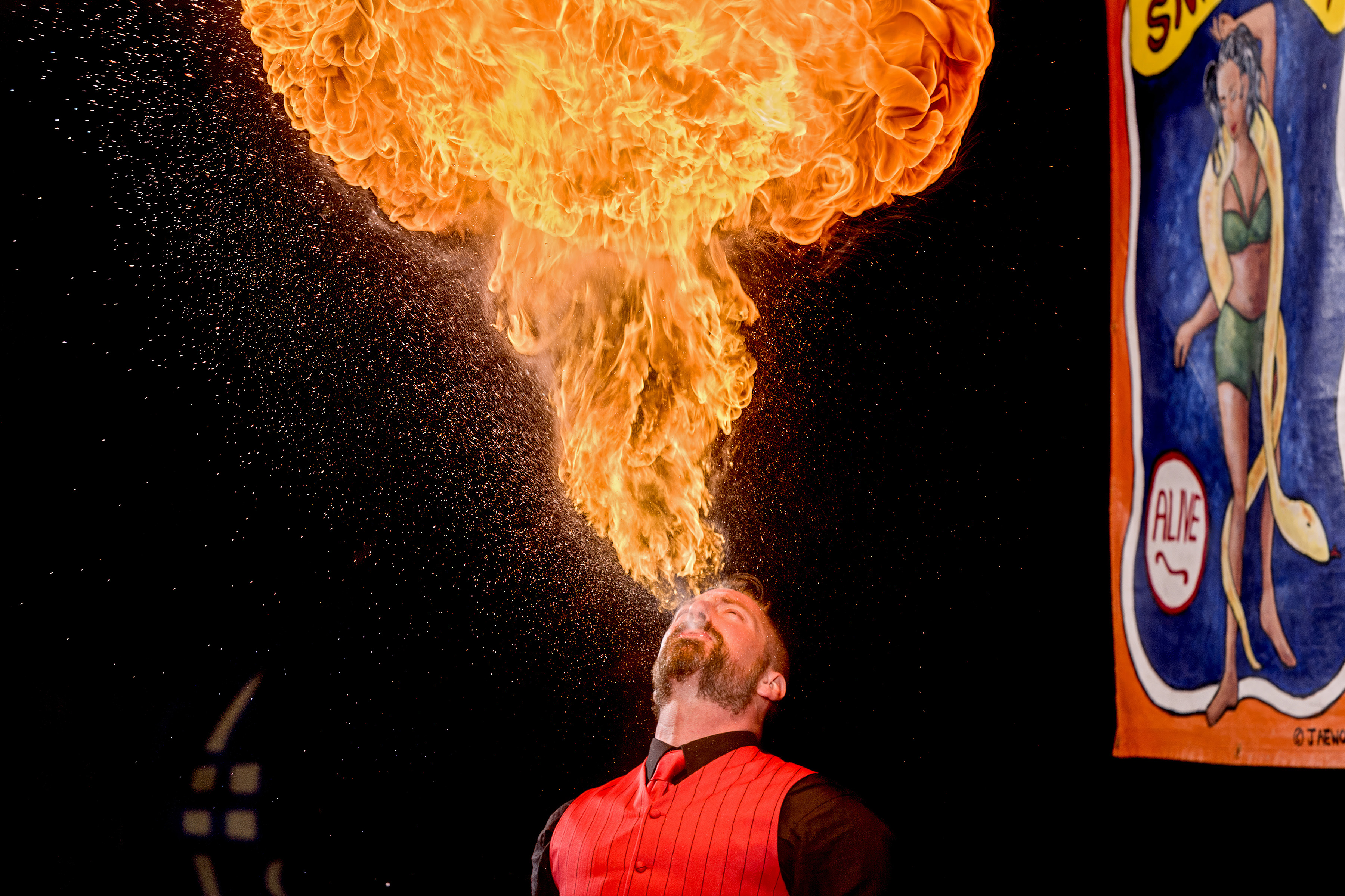 Man in red shirt breathing large orange flame upwards against black background, with colourful circus poster visible on right.