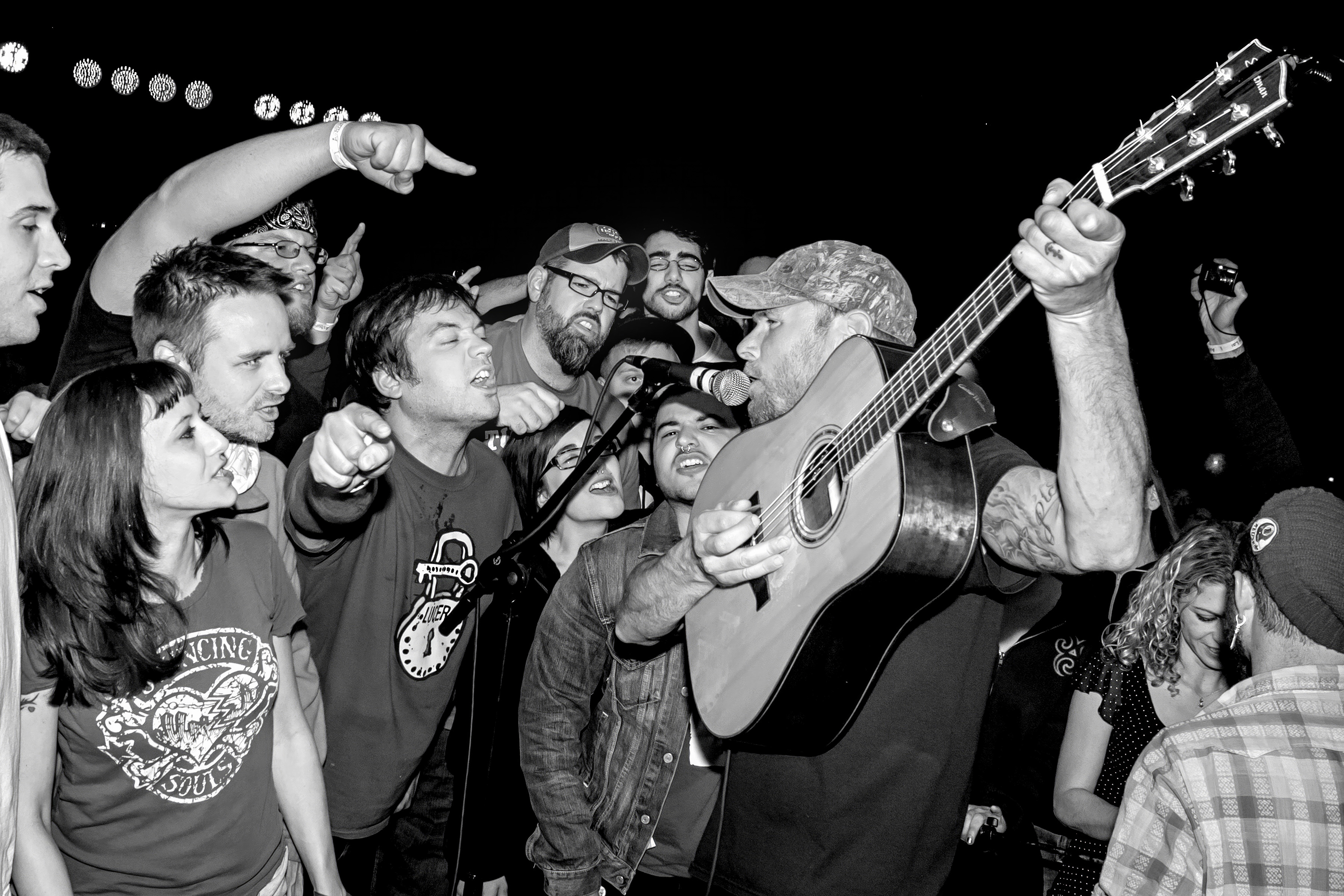 Black and white image of guitarist performing surrounded by enthusiastic crowd, acoustic guitar raised above heads, audience reaching towards performer.