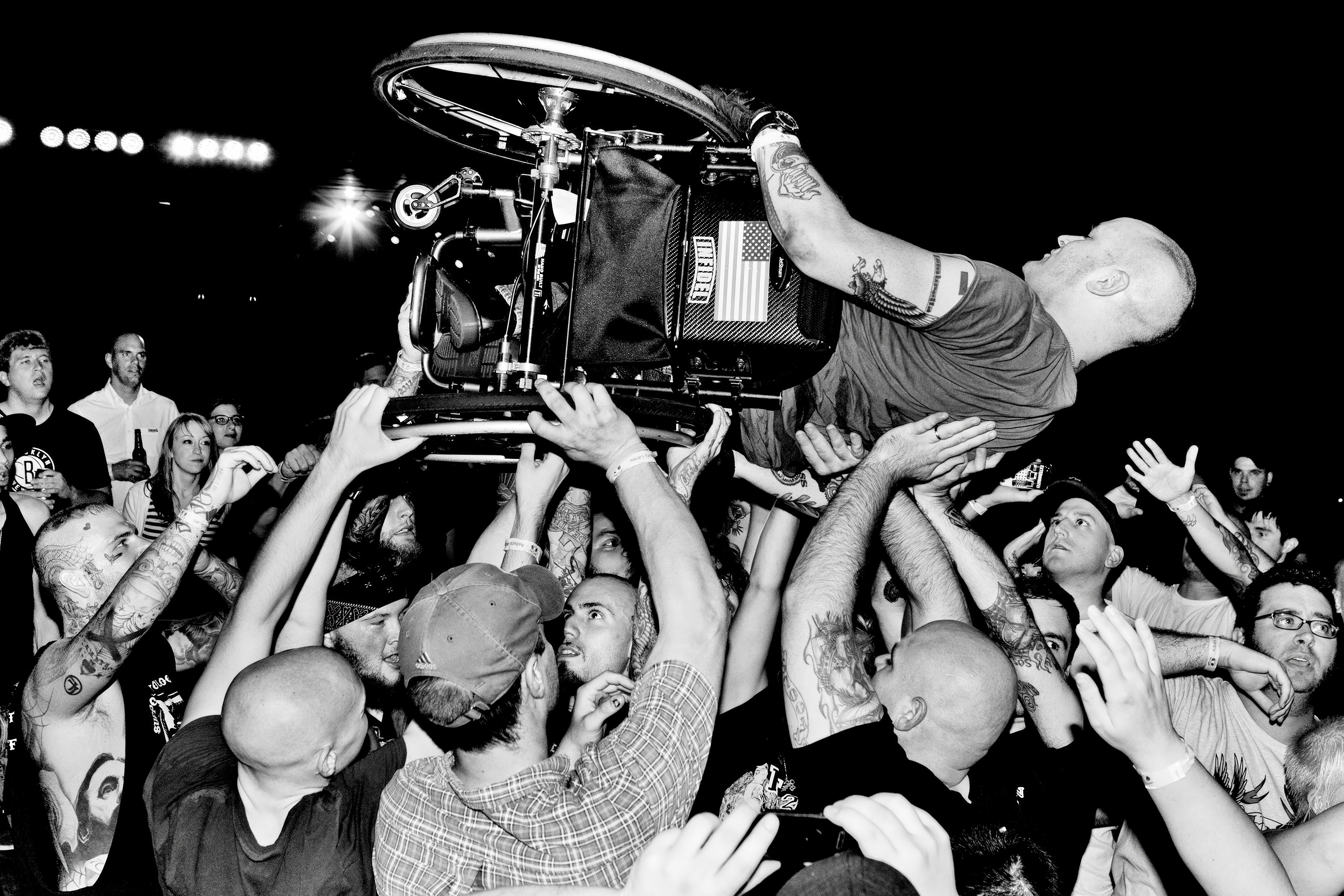 Black and white image of performer crowd-surfing with wheelchair above concert audience, stage lights visible in background.