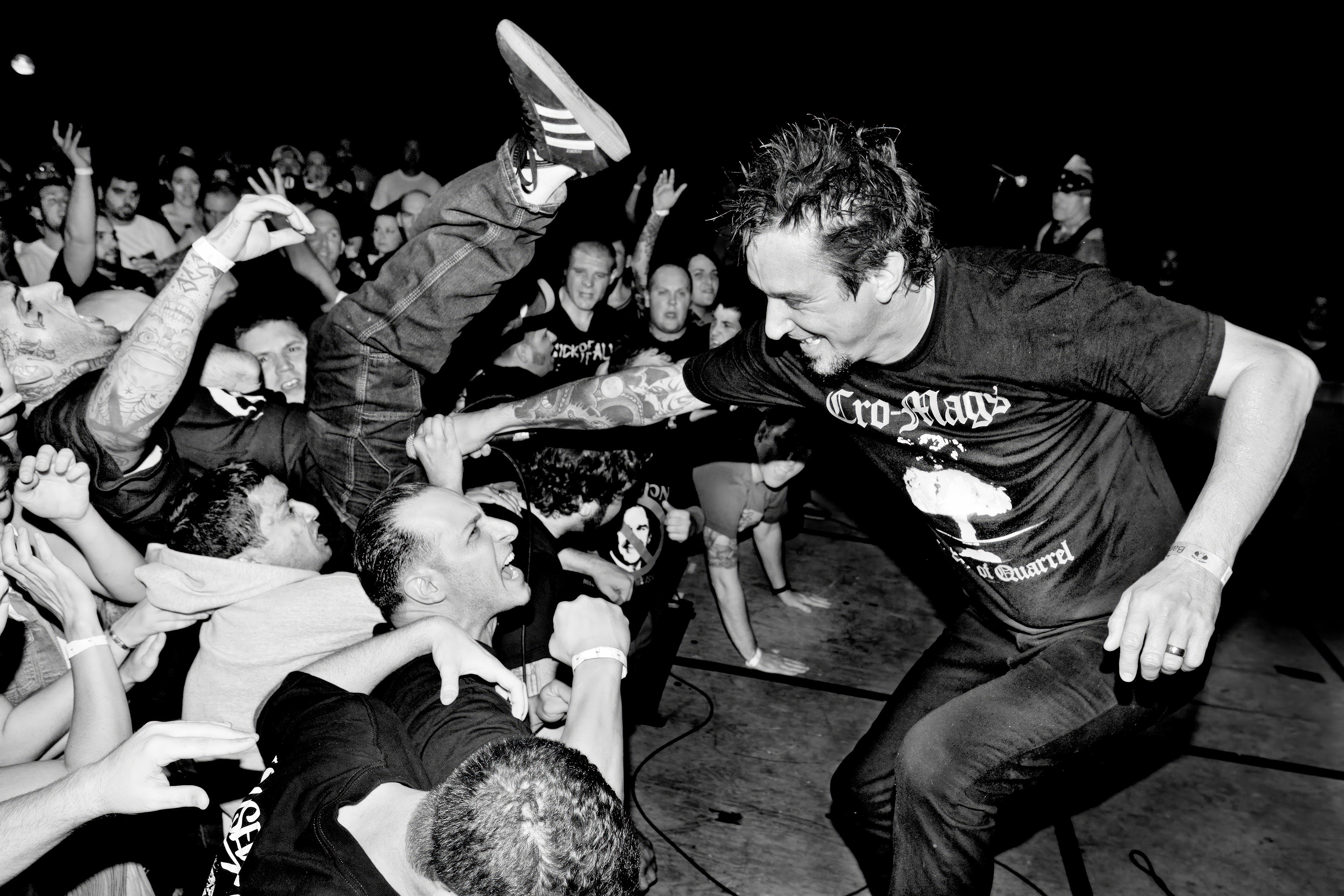 Black and white image of performer in dark t-shirt leaning towards seated crowd, reaching out to audience members in concert venue.