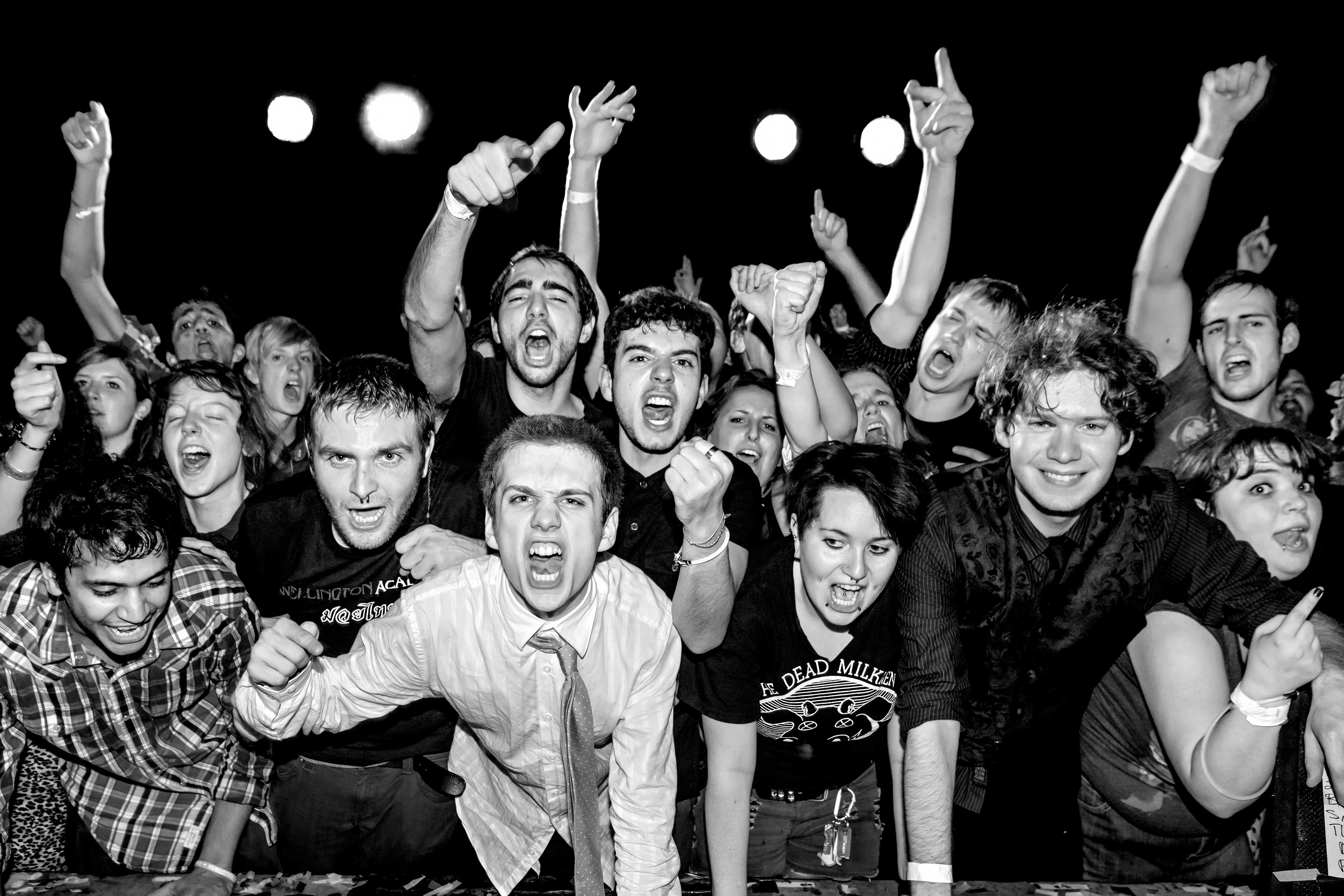 Black and white image of crowded concert audience with raised fists and arms, bright stage lights visible in background.