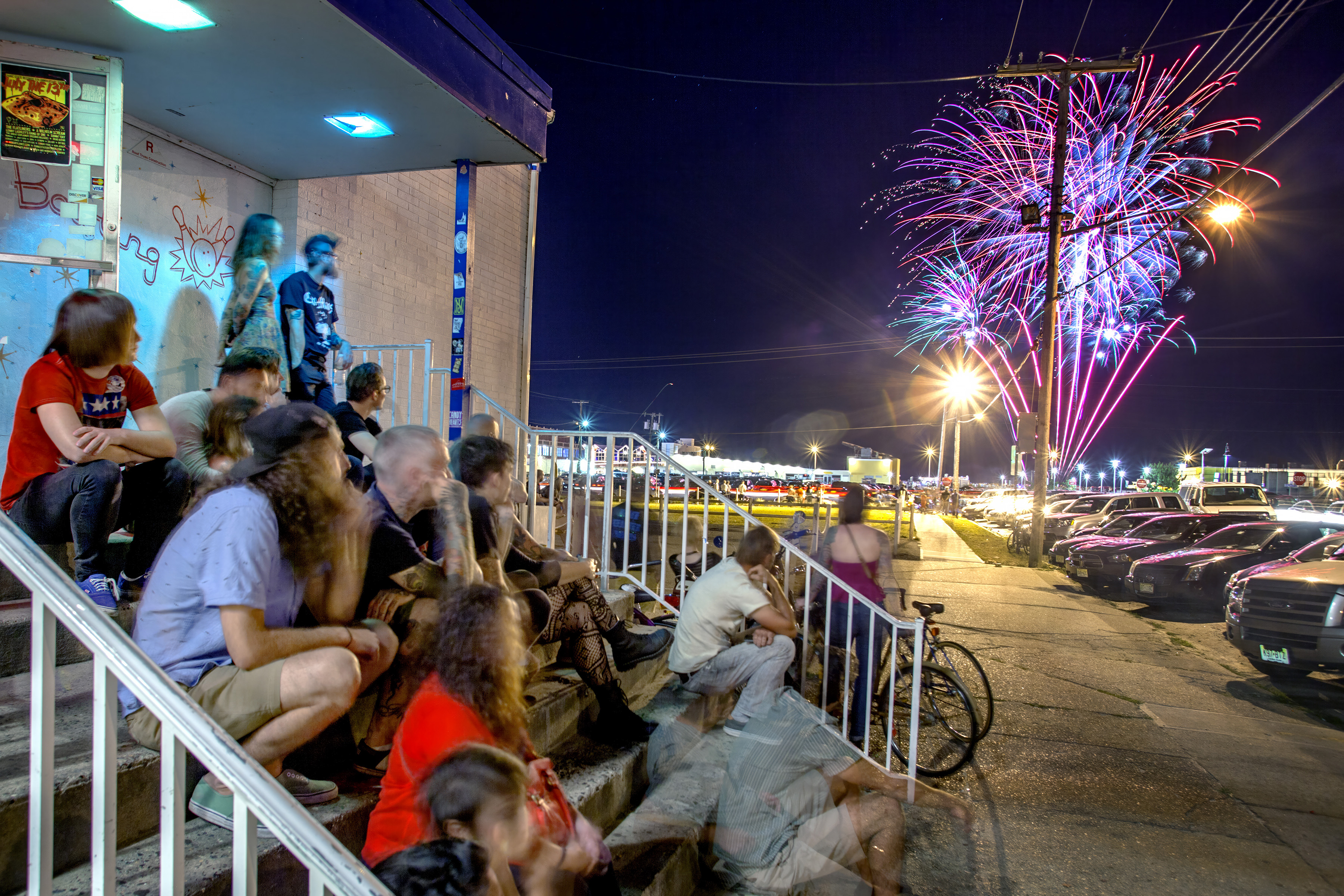People sitting on tiered seating watching colourful fireworks display over harbour at night with parked cars and street lights visible.