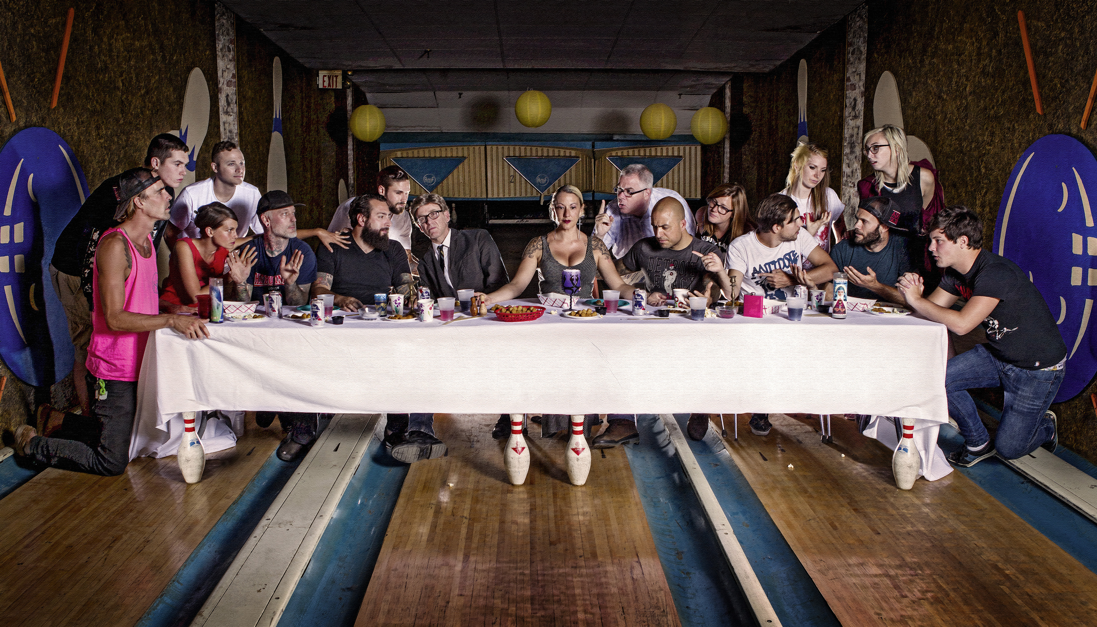 Large group seated around white-clothed table in bowling alley, with lanes and pins visible, recreating Last Supper composition.