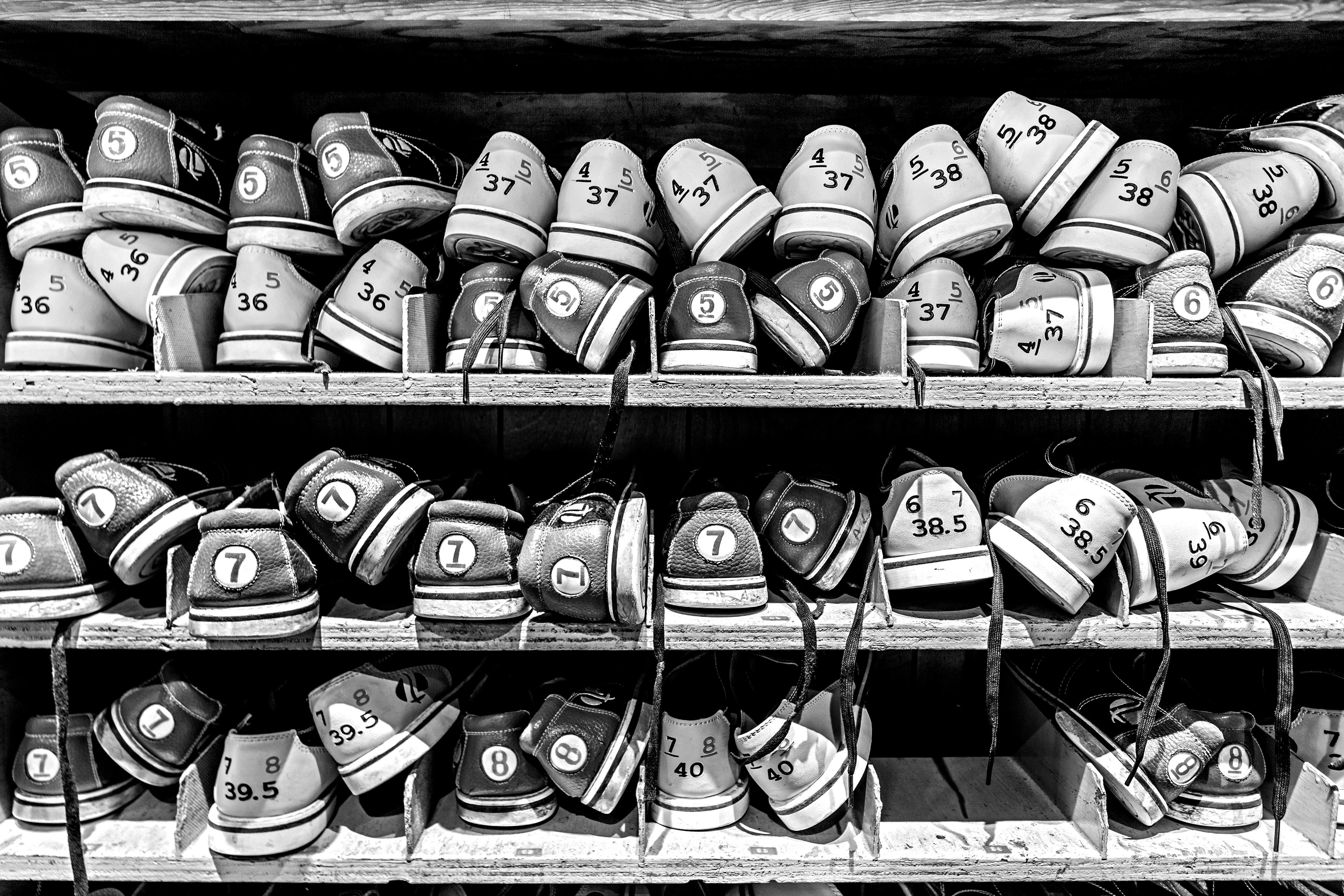 Black and white image showing three shelves filled with numbered bowling shoes arranged in compartments, sizes visible on labels.