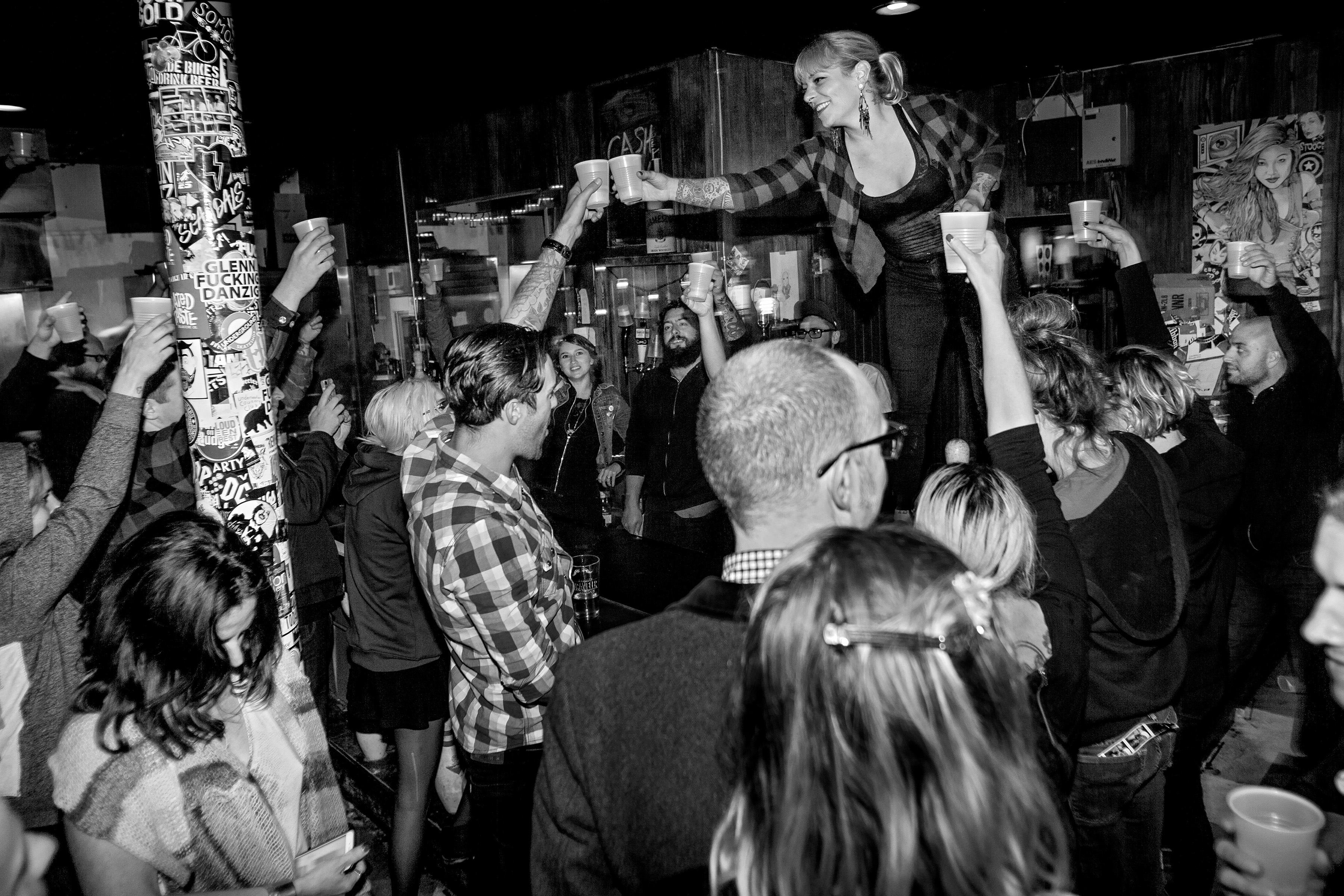Black and white image of crowded indoor venue with person standing elevated amongst audience, arms outstretched holding drinks.