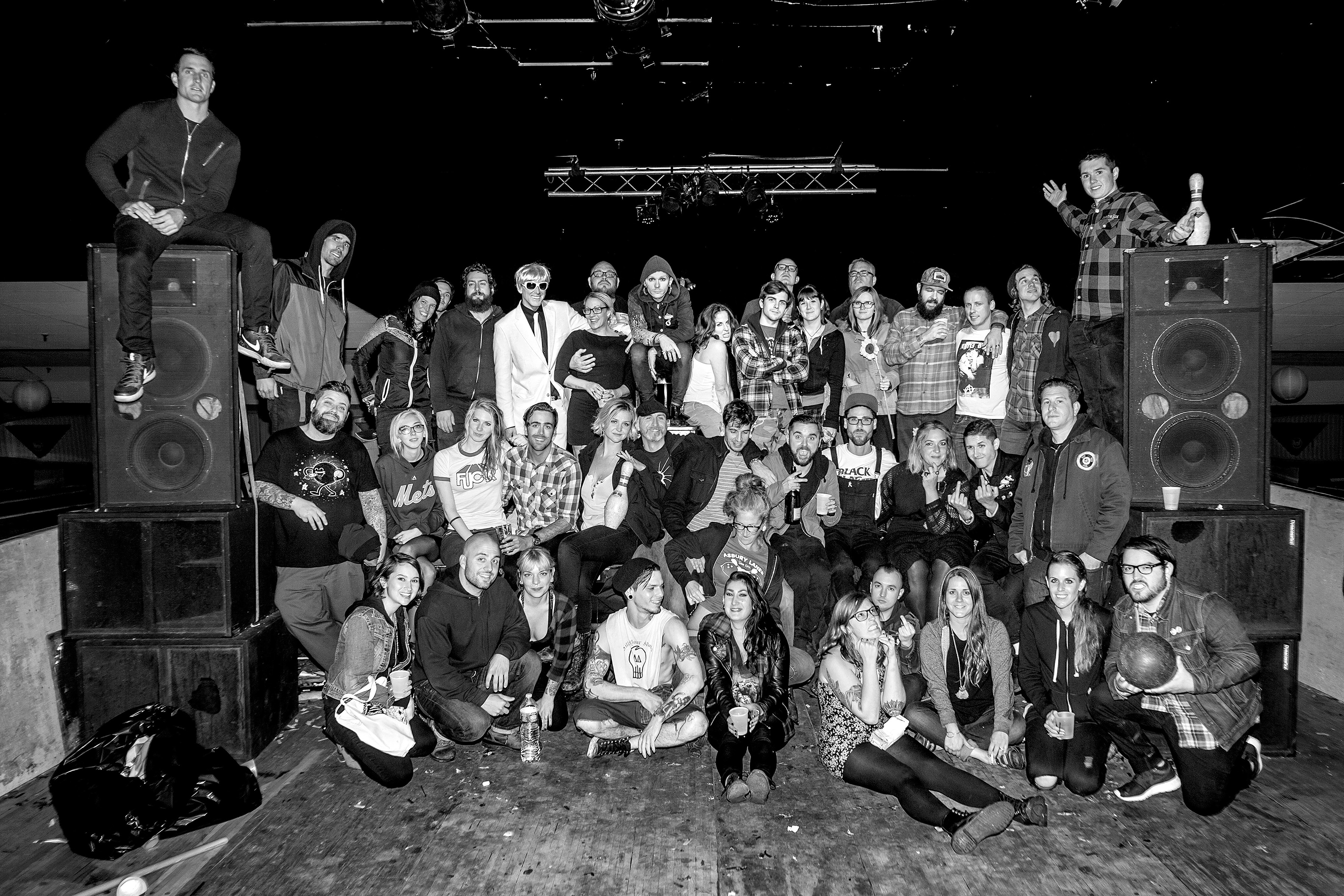 Large group of people posed together in black and white, surrounded by stacked speakers and sound equipment in what appears to be a venue or warehouse space.