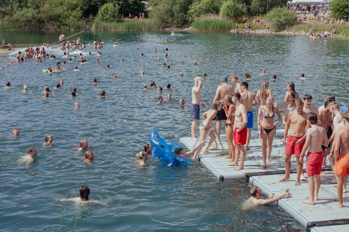 Crowded lake with swimmers in water and people standing on wooden jetty, surrounded by green trees and distant buildings.
