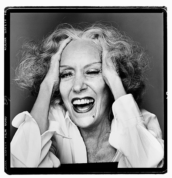 Black and white portrait of woman with curly hair laughing, hands framing face, wearing light-coloured shirt. Film negative border visible.