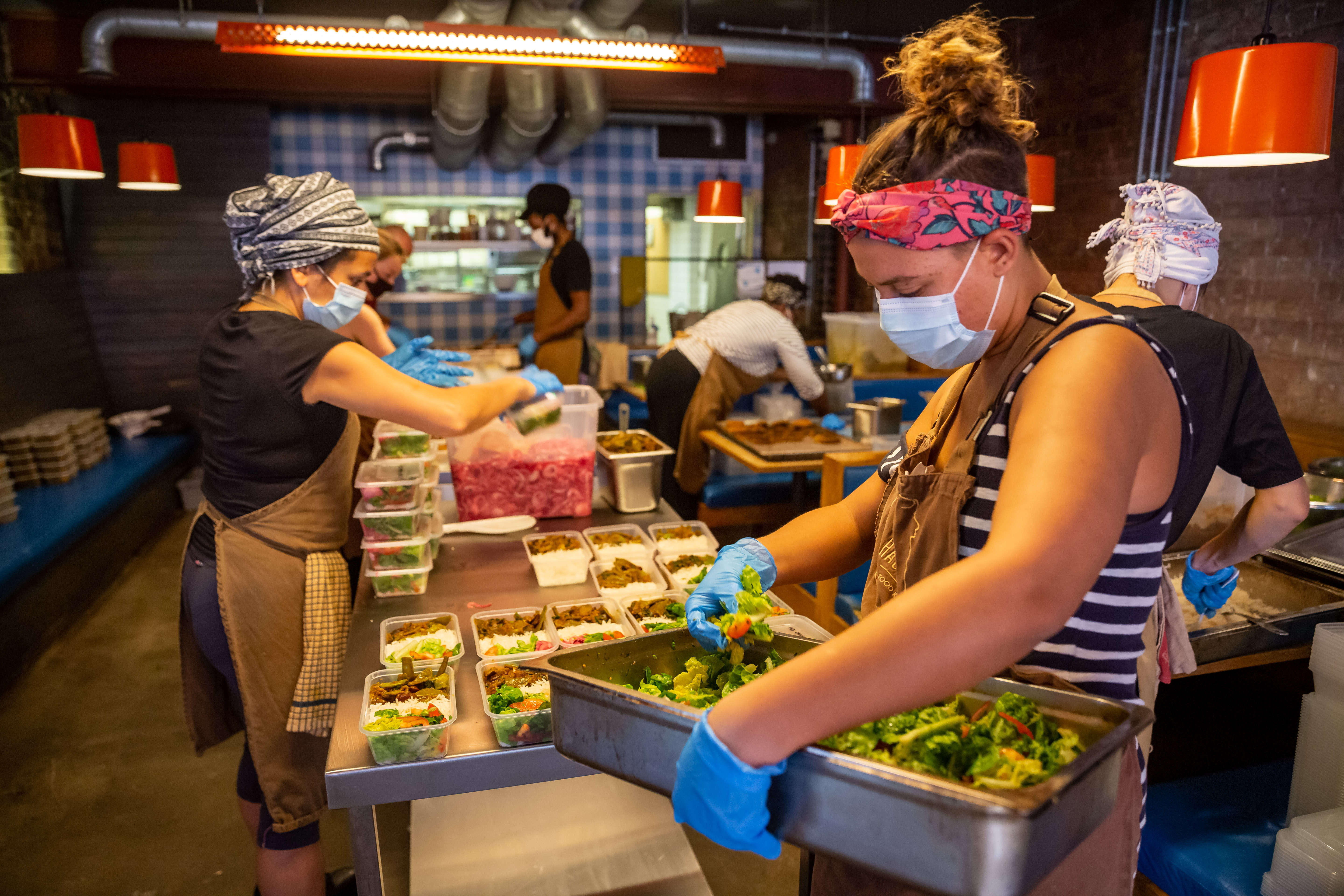 Kitchen staff wearing masks and hairnets prepare food under heat lamps, with one woman plating salad in foreground wearing striped apron.
