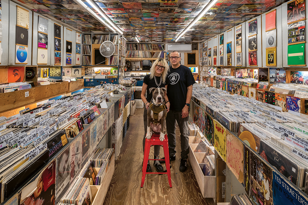 Two people and a dog on red stool stand in record shop aisle surrounded by vinyl records covering walls and ceiling.