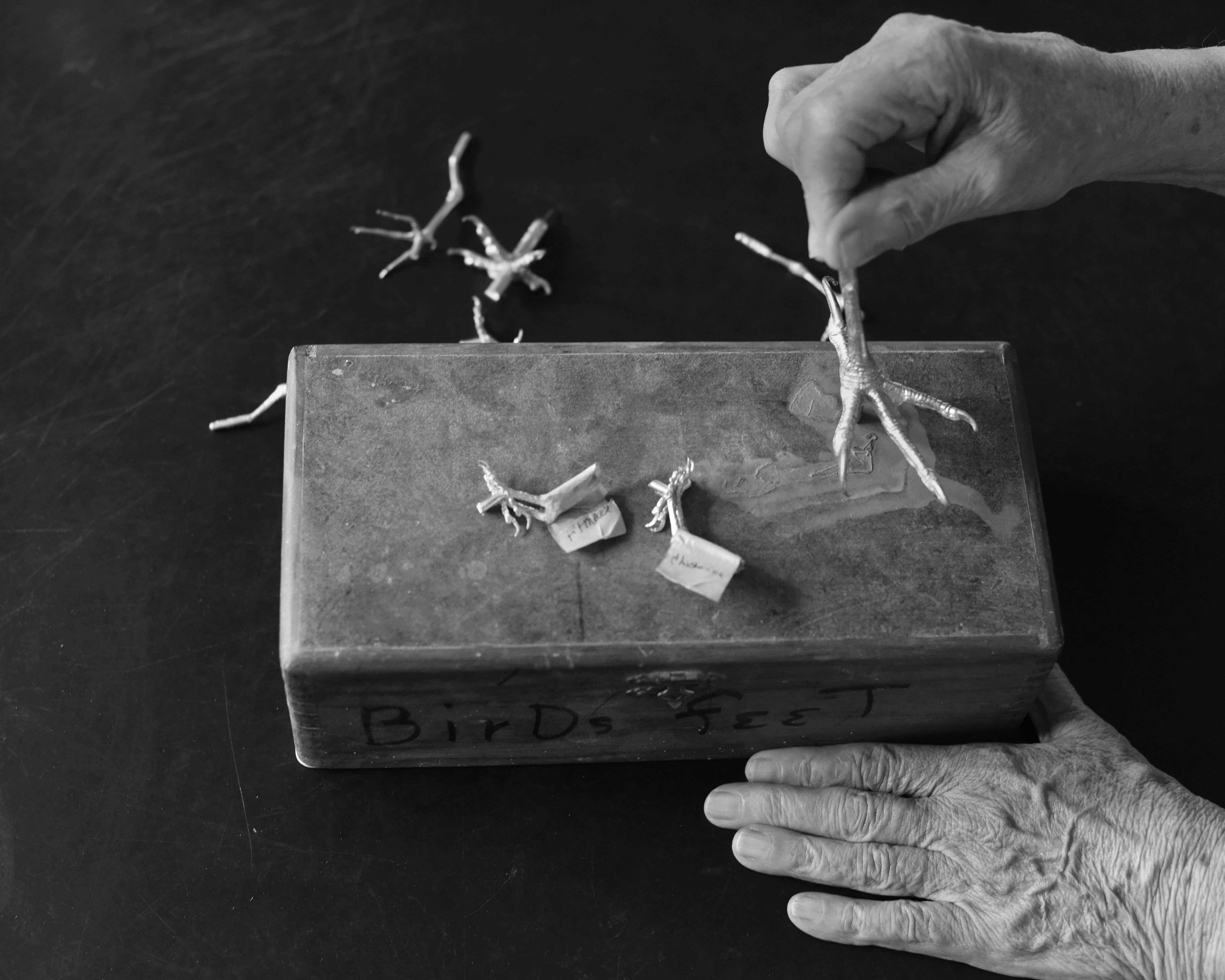 Black and white image showing hands manipulating small figurines on a wooden box surface against dark background.