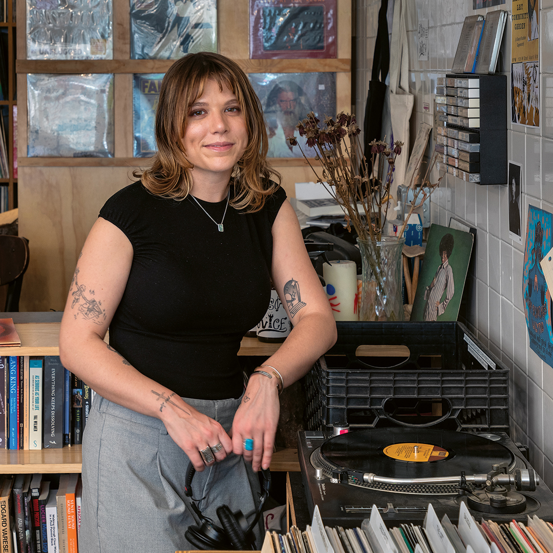 Woman with shoulder-length blonde hair and tattoos sits in art studio wearing black top, surrounded by books, artwork, and creative materials.