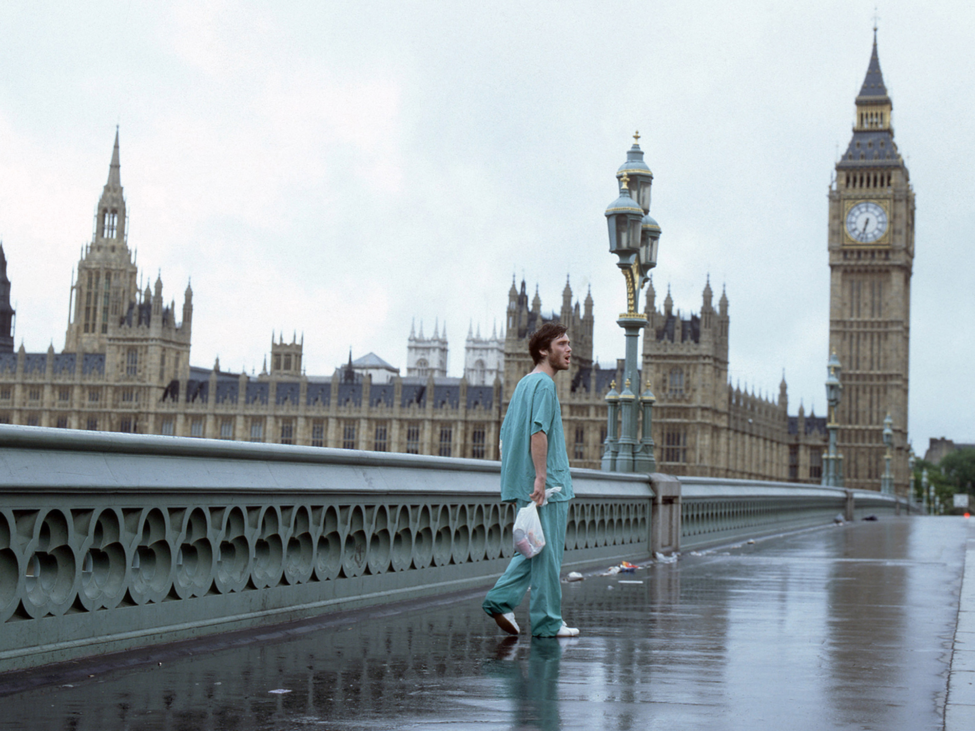 Cilliam Murphy in green hospital scrubs walking on a deserted Westminster Bridge with Houses of Parliament and Big Ben in background.