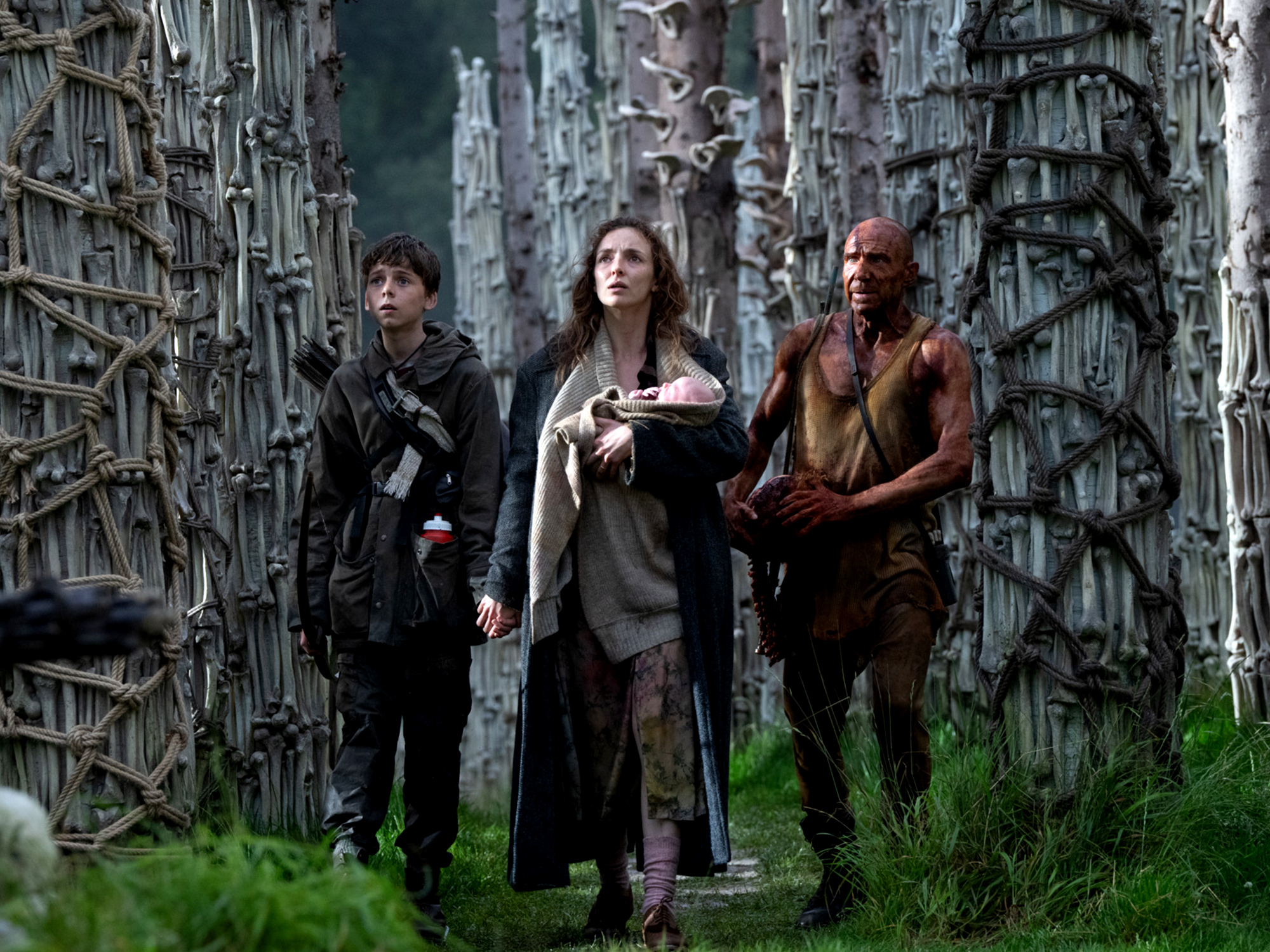 Three people walking through a dense, overgrown landscape surrounded by tangled branches and foliage.