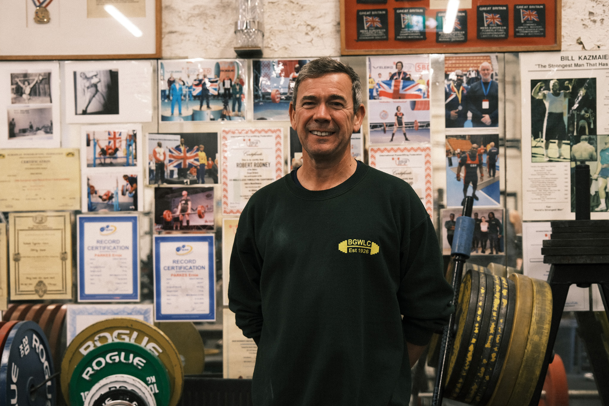 Man in green jumper standing in workshop with wall display of certificates, photographs, and memorabilia behind him.
