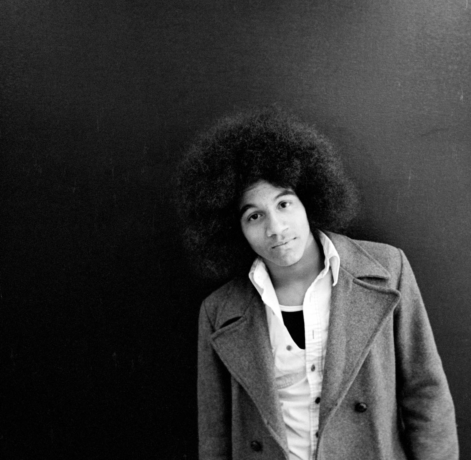 Black and white portrait of person with large afro hairstyle wearing light-coloured coat, positioned right of frame against dark background.