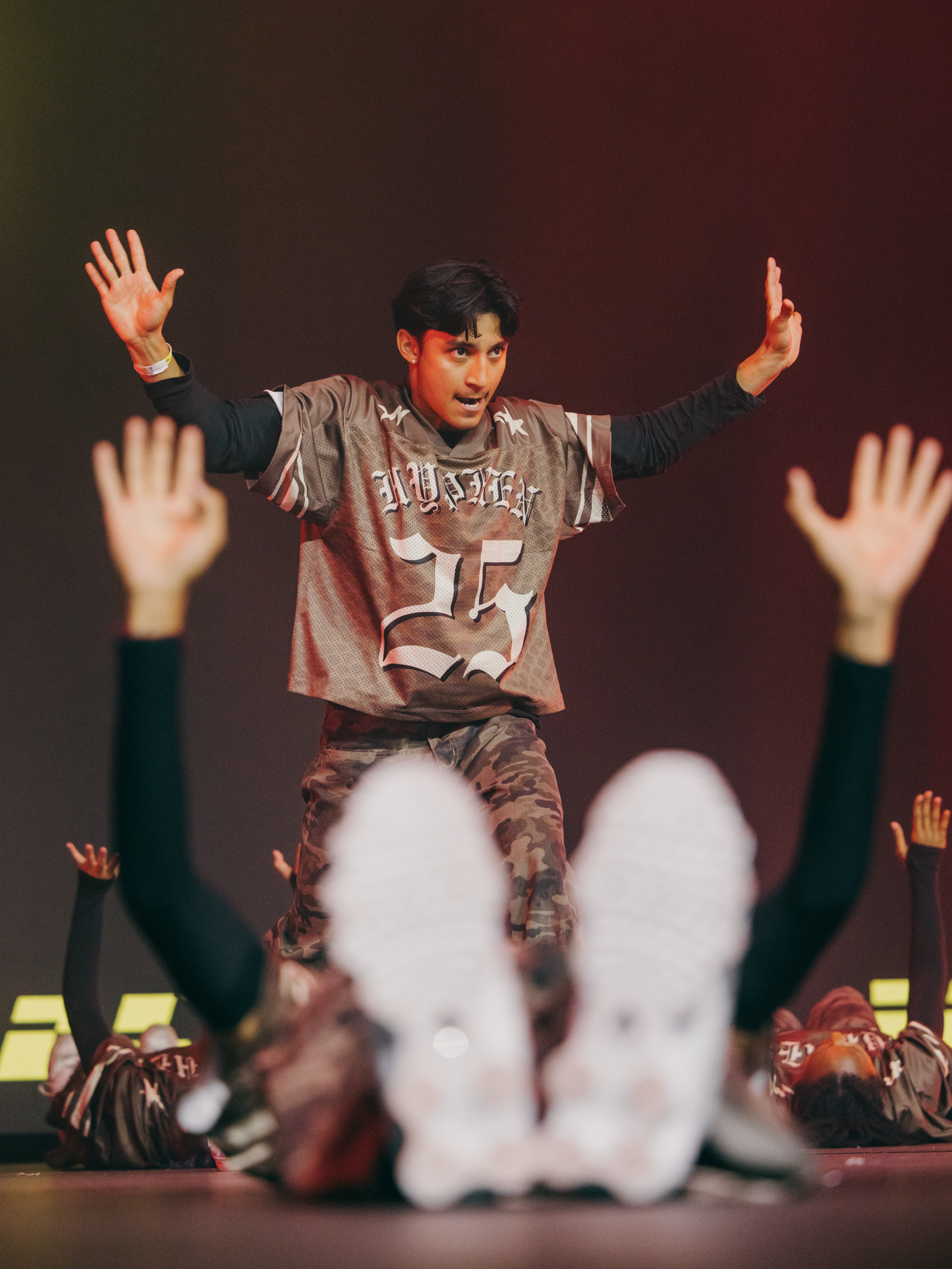 Performer in grey sports jersey with raised arms on stage, audience members' hands raised in foreground, dark red background lighting.
