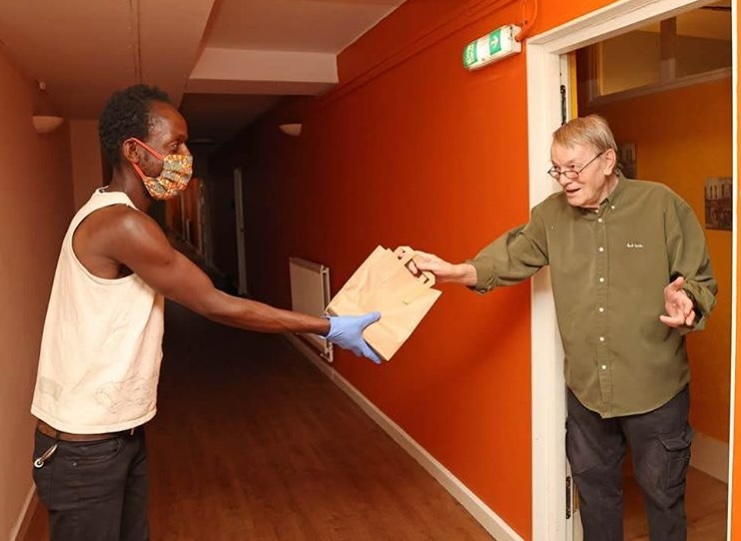 Two men exchanging an envelope in orange-walled corridor. One wears white vest and mask, other in green shirt and glasses.