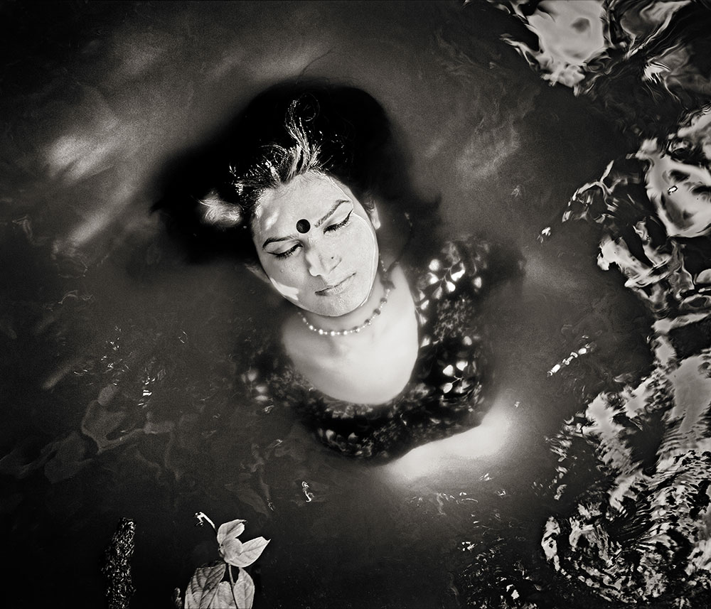Woman floating in dark water wearing pearls, surrounded by murky greenish-brown tones with scattered white reflective elements.