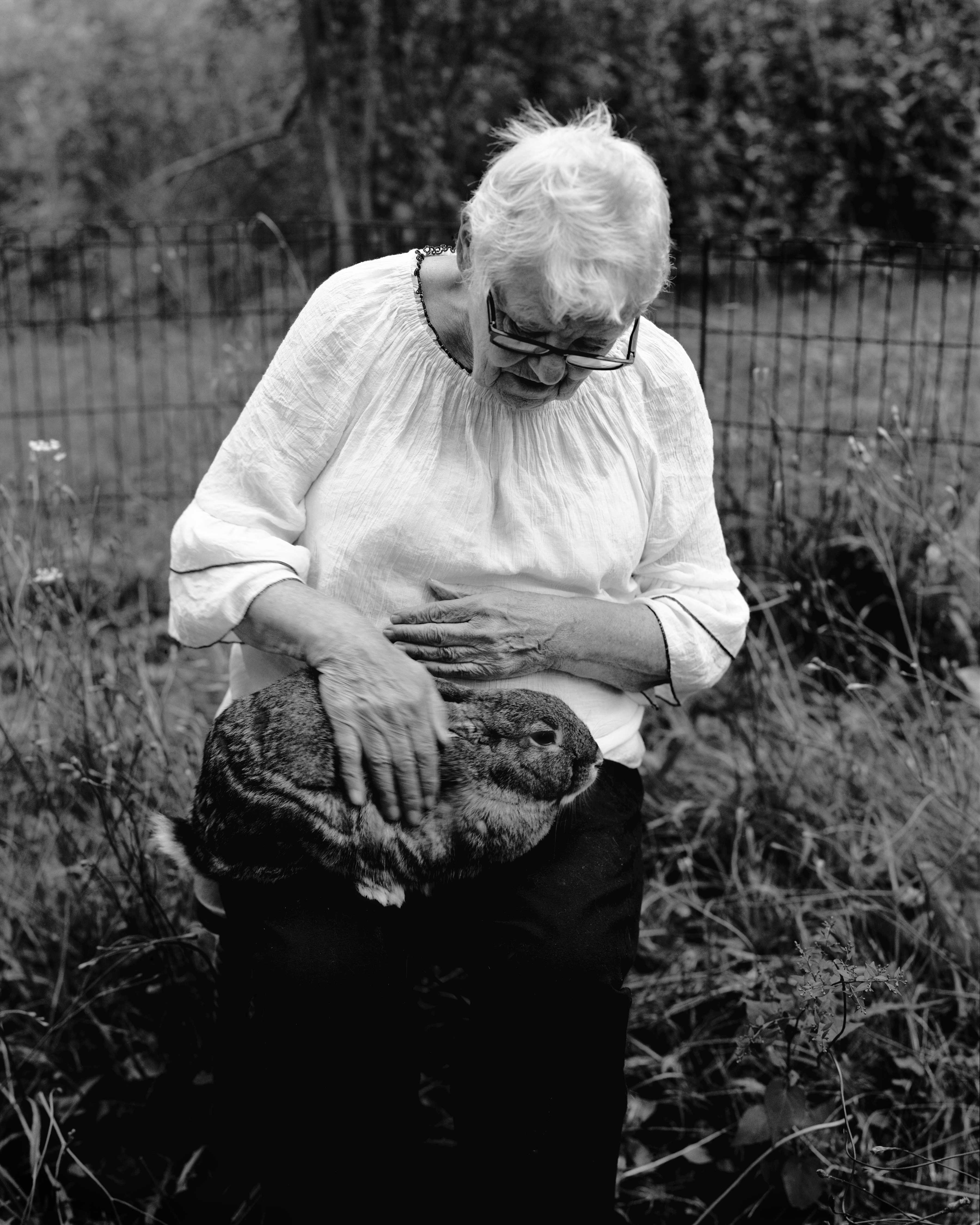 Elderly woman with glasses and white hair kneeling outdoors, petting a rabbit on her lap. Black and white photograph.