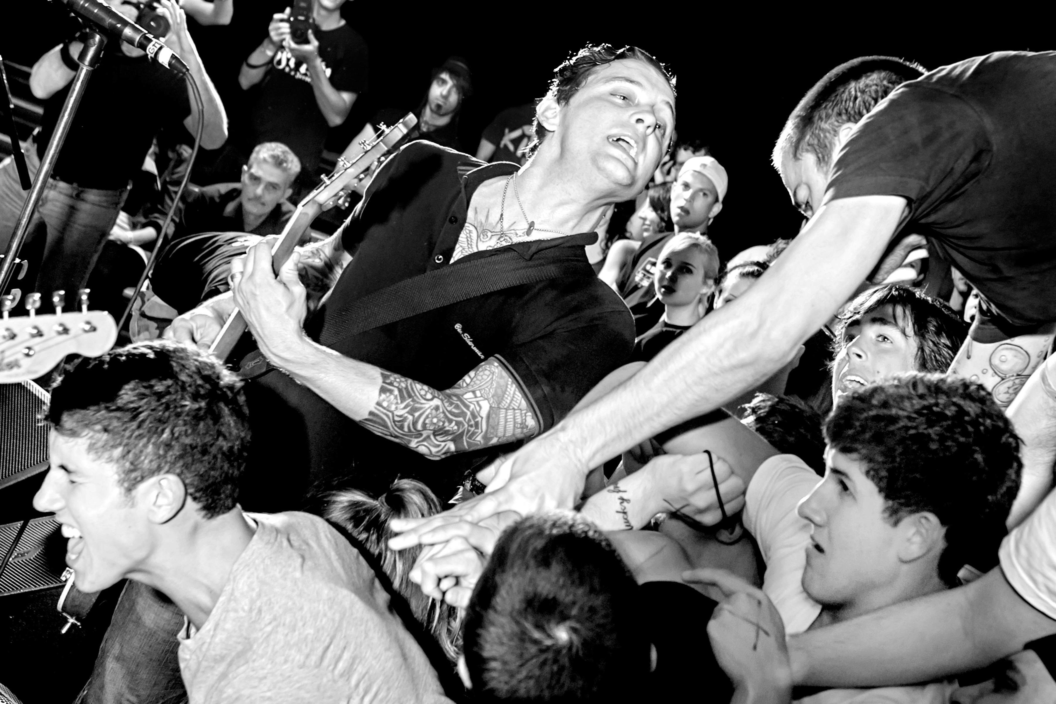Black and white image of performer crowd-surfing at concert, surrounded by reaching hands and faces looking upward.