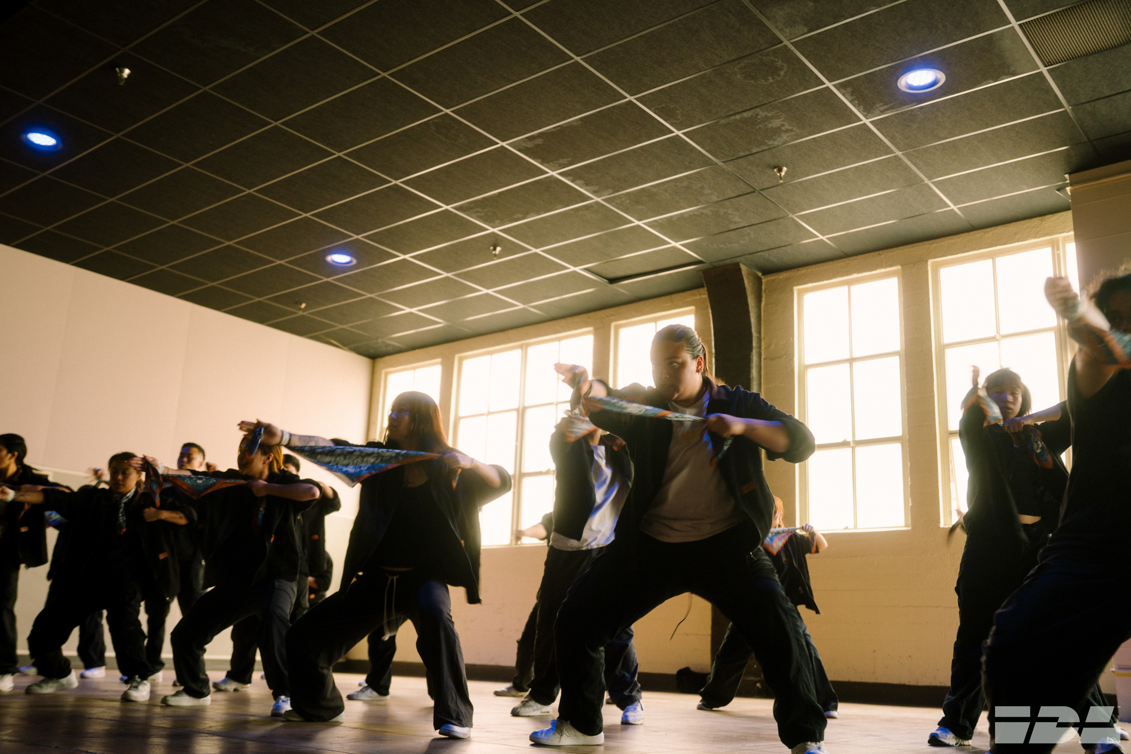 Group of people in black clothing practising martial arts movements in indoor studio with large windows and tiled ceiling.