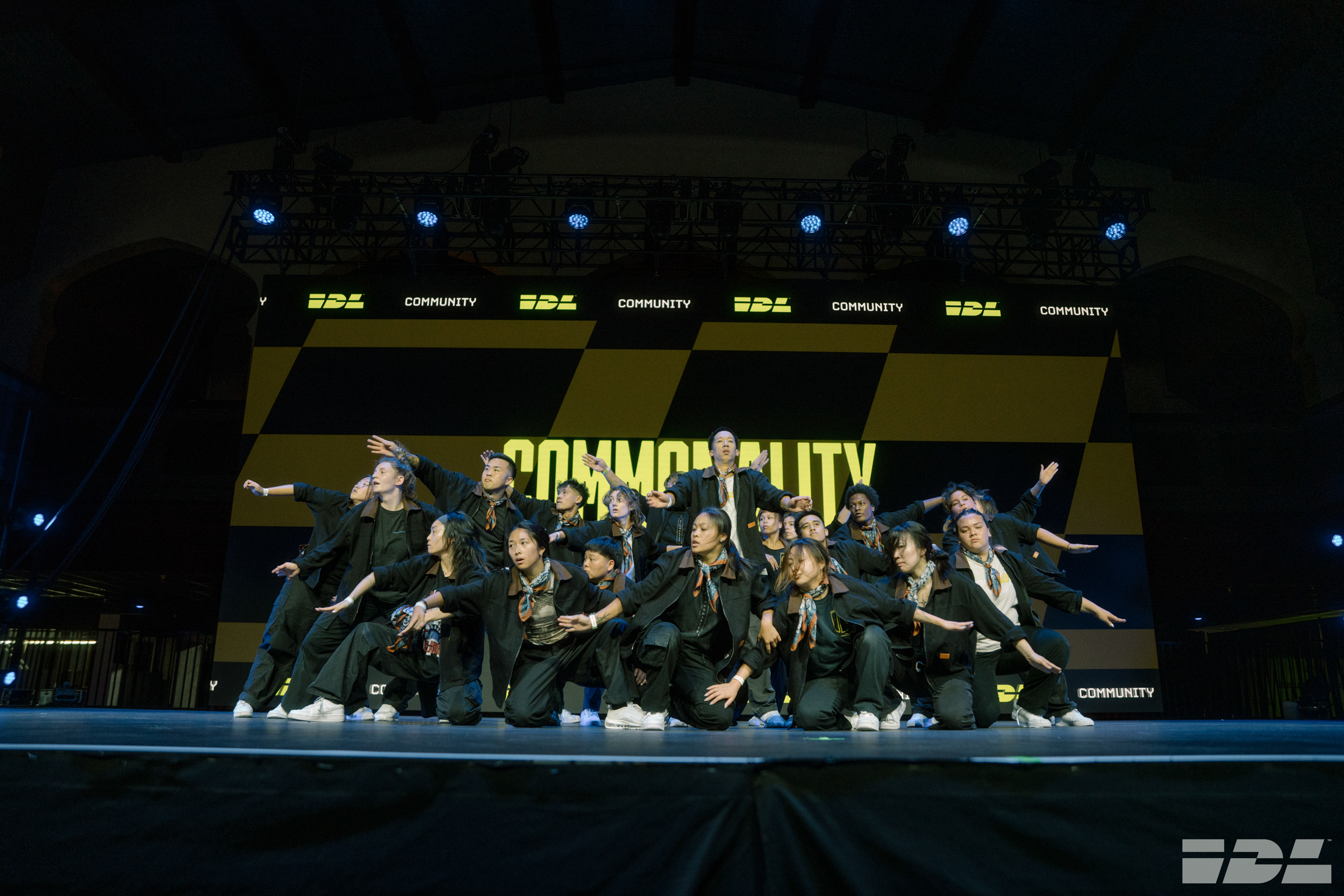 Dance group in black outfits performing on stage with yellow "COMMUNITY" backdrop and blue lighting.