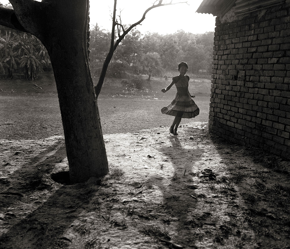 Child in dress dancing between tree and brick building on muddy ground in misty outdoor setting.
