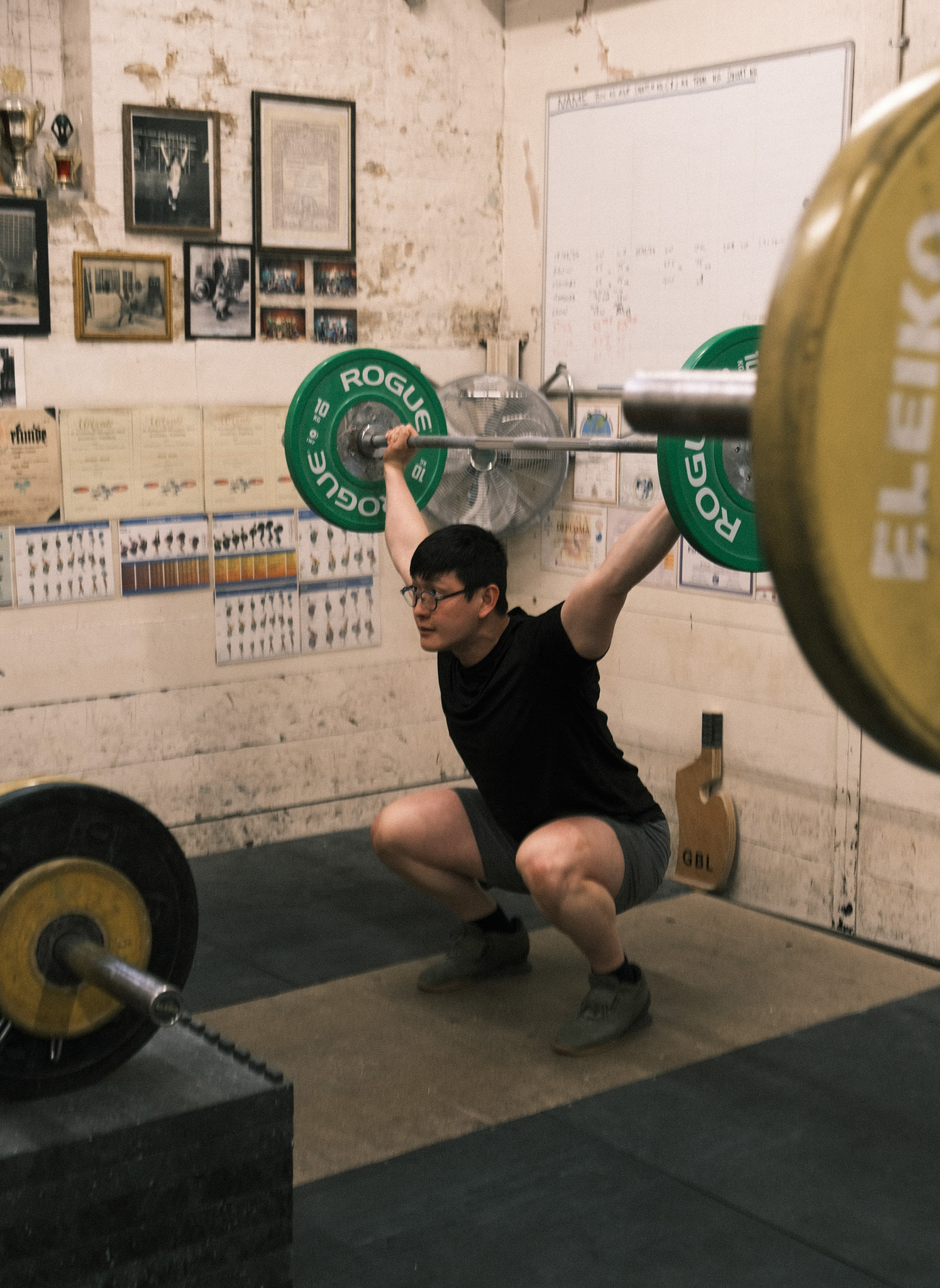 Man in black shirt performing overhead squat with barbell and green Rogue weight plates in gym with white brick walls.