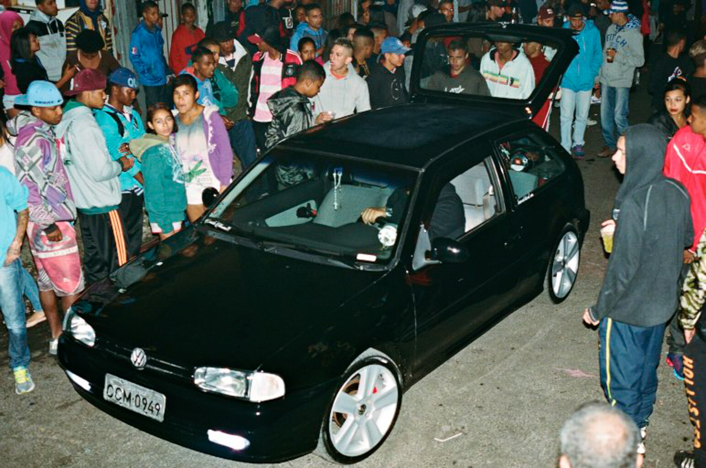 Black Volkswagen Golf with white alloy wheels surrounded by crowd of people on street at night.
