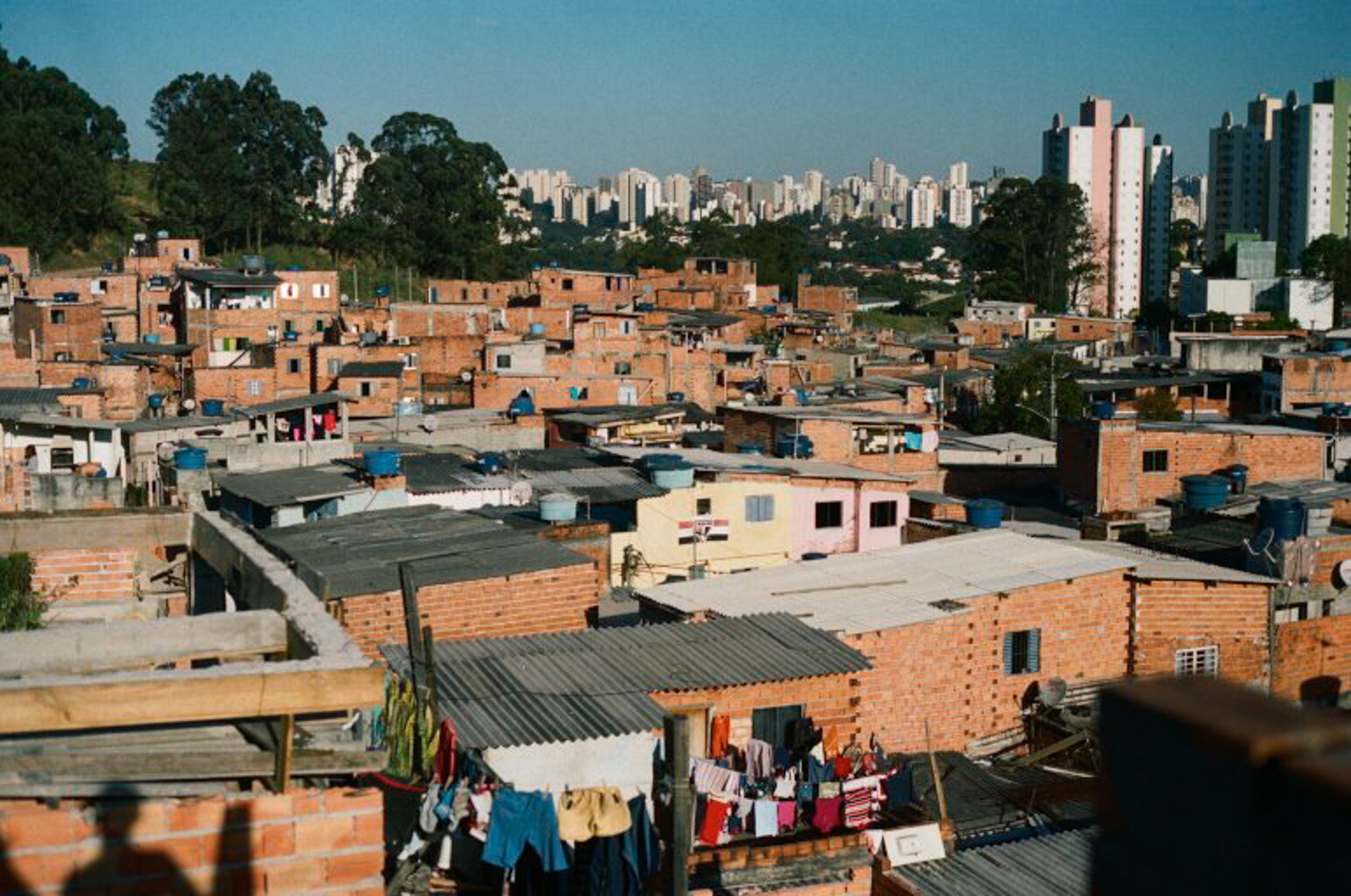 Dense informal settlement with orange brick buildings and metal roofs in foreground, modern city skyline visible in background.