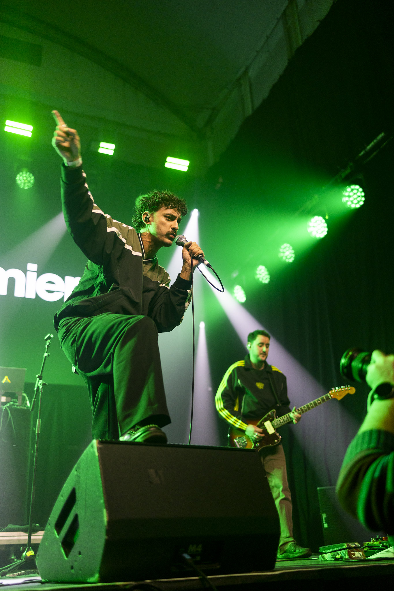 Singer pointing upward with microphone on stage, guitarist in background, green stage lighting and white spotlights, audience visible.