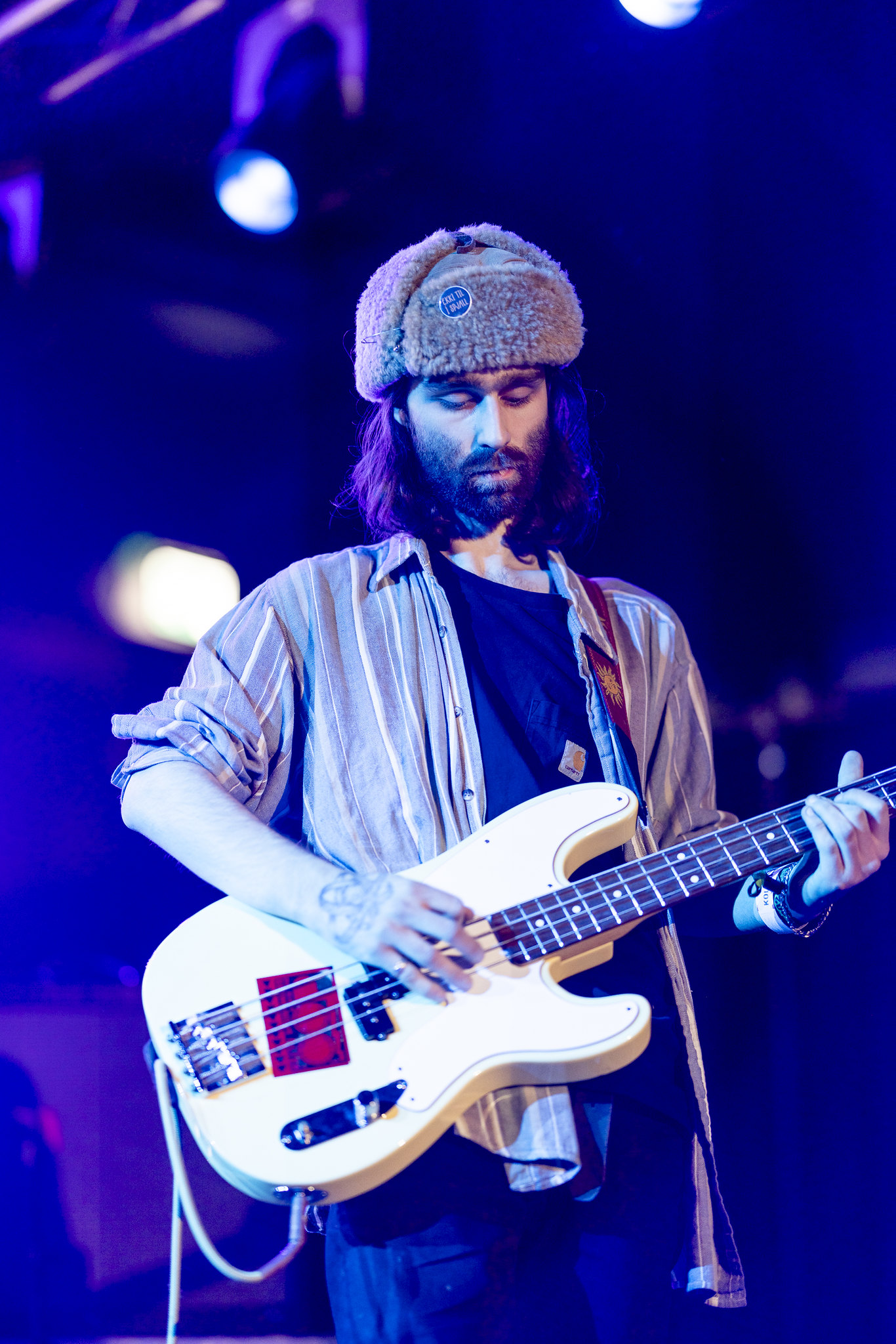 Bearded musician wearing knit hat playing white bass guitar on stage under purple and blue stage lighting.