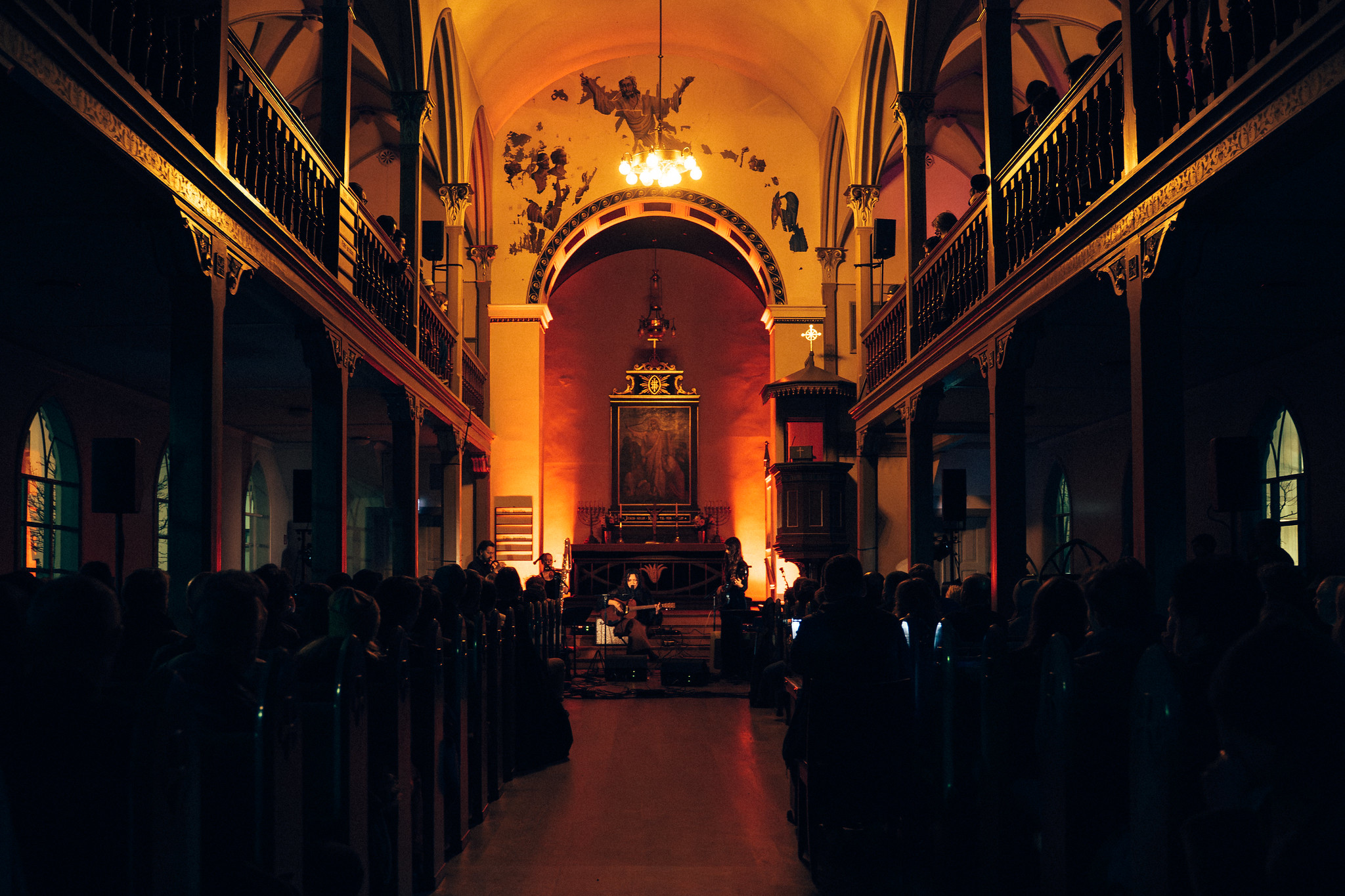 Church interior with orange lighting illuminating arched ceiling and altar area, dark wooden pews in foreground, balconies on sides.