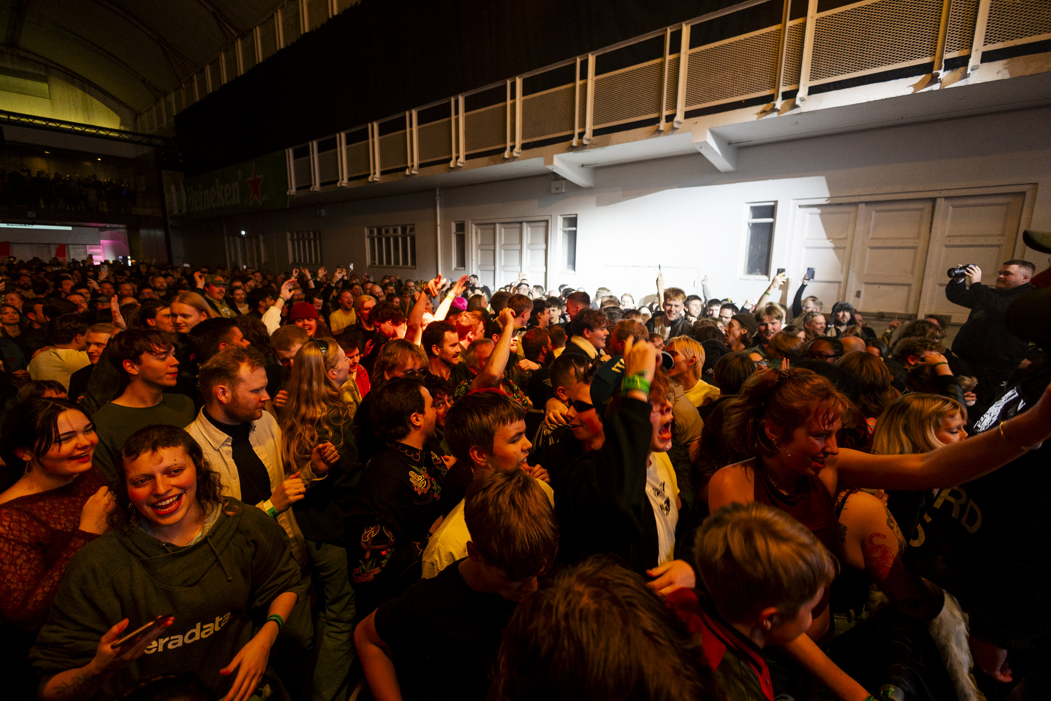 Large crowd gathered in courtyard at night, illuminated by warm orange lighting, with white colonial-style building featuring balcony in background.