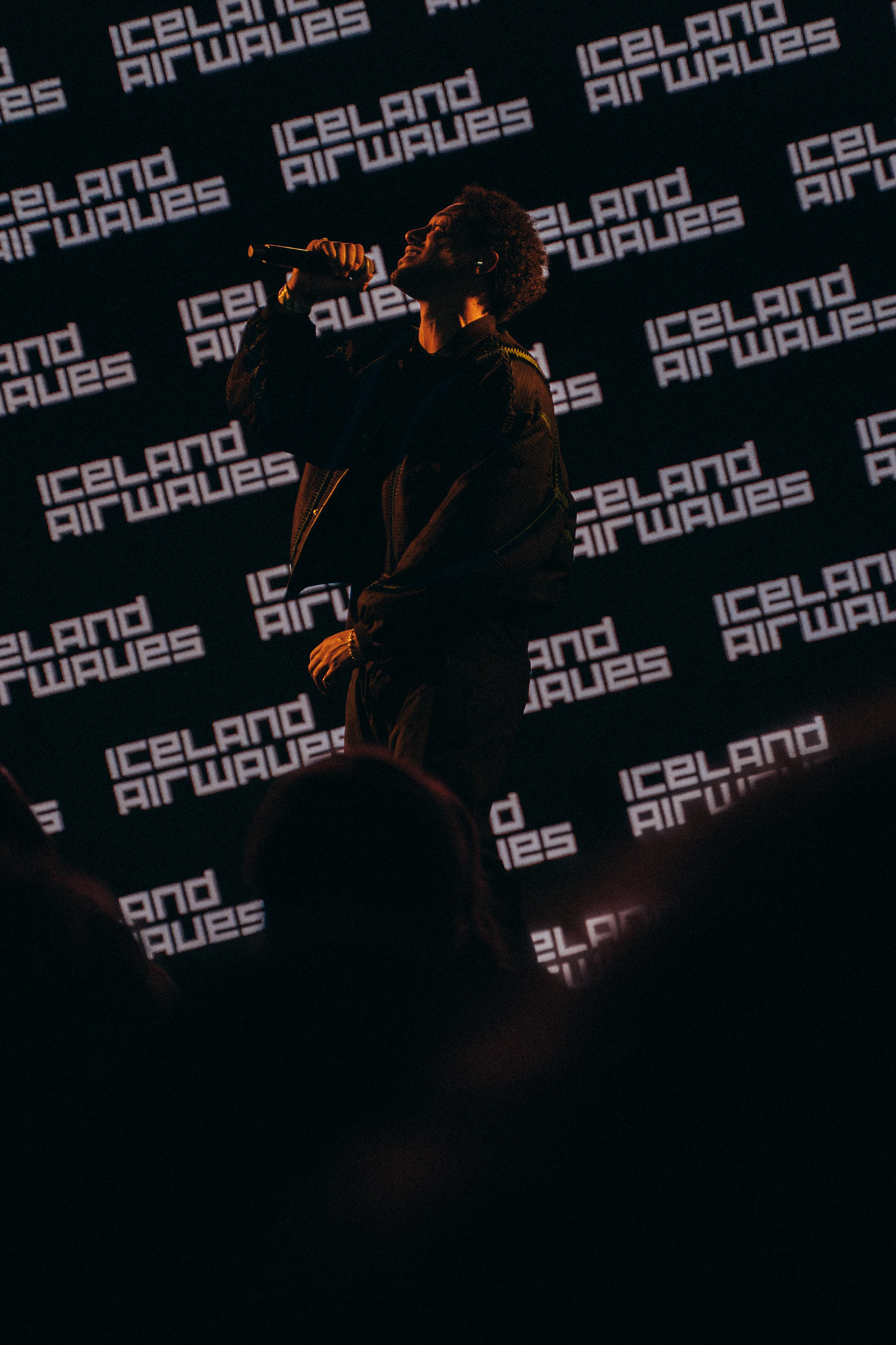 Person in dark clothing with orange lighting accents standing against black backdrop featuring repeated "Iceland Airwaves" text logos.