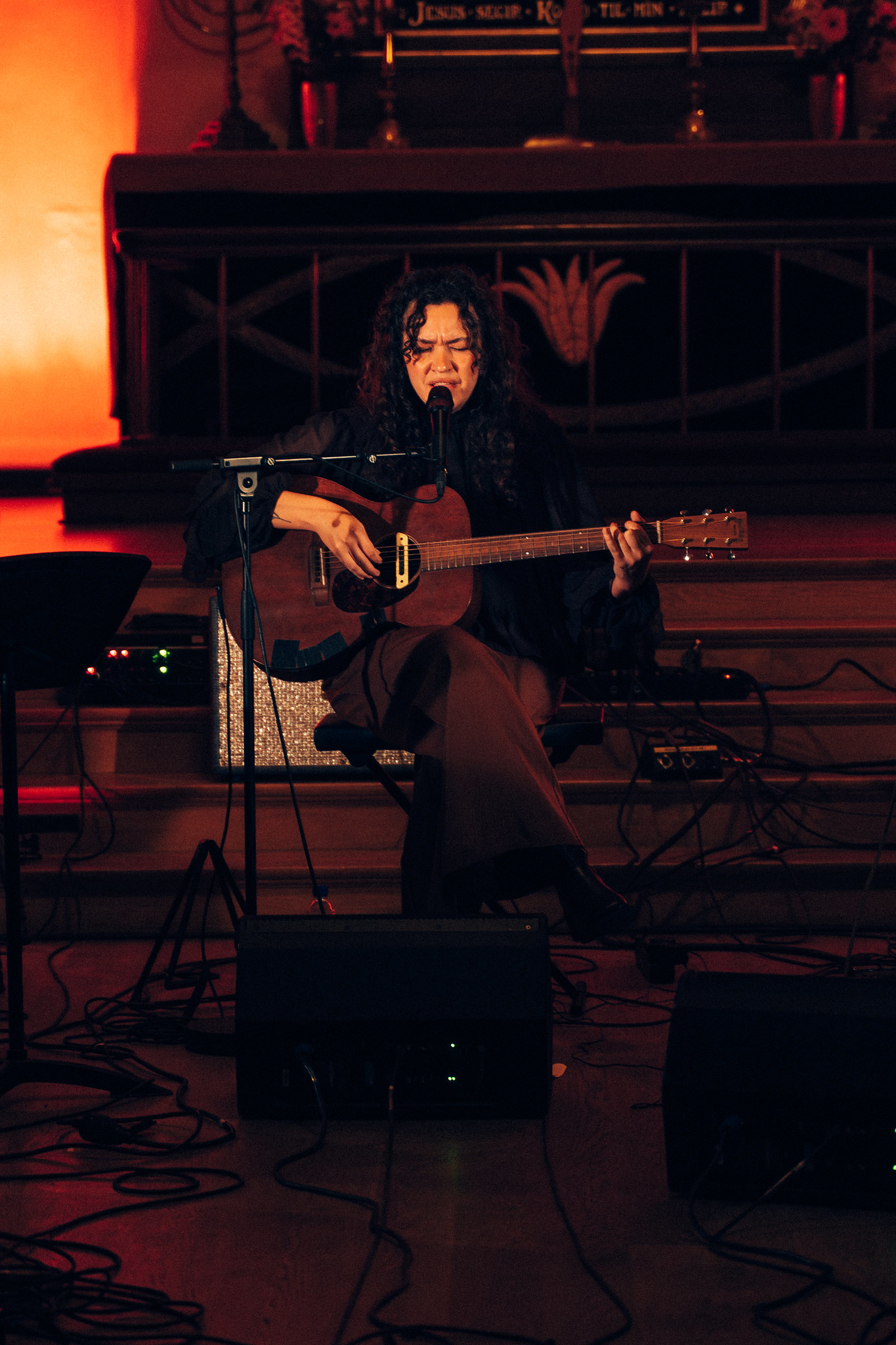 Man with long dark hair playing acoustic guitar and singing into microphone on dimly lit stage with warm orange lighting.
