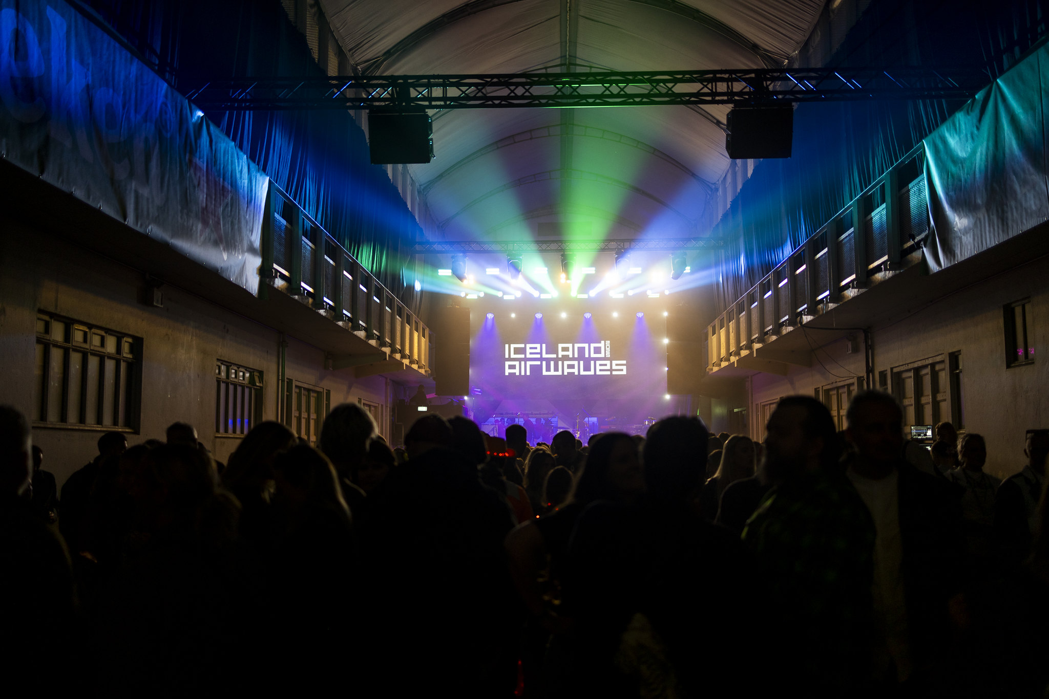 Concert venue with silhouetted crowd facing illuminated stage. Green and blue lights beam from overhead, text on screen reads "Iceland Airwaves".
