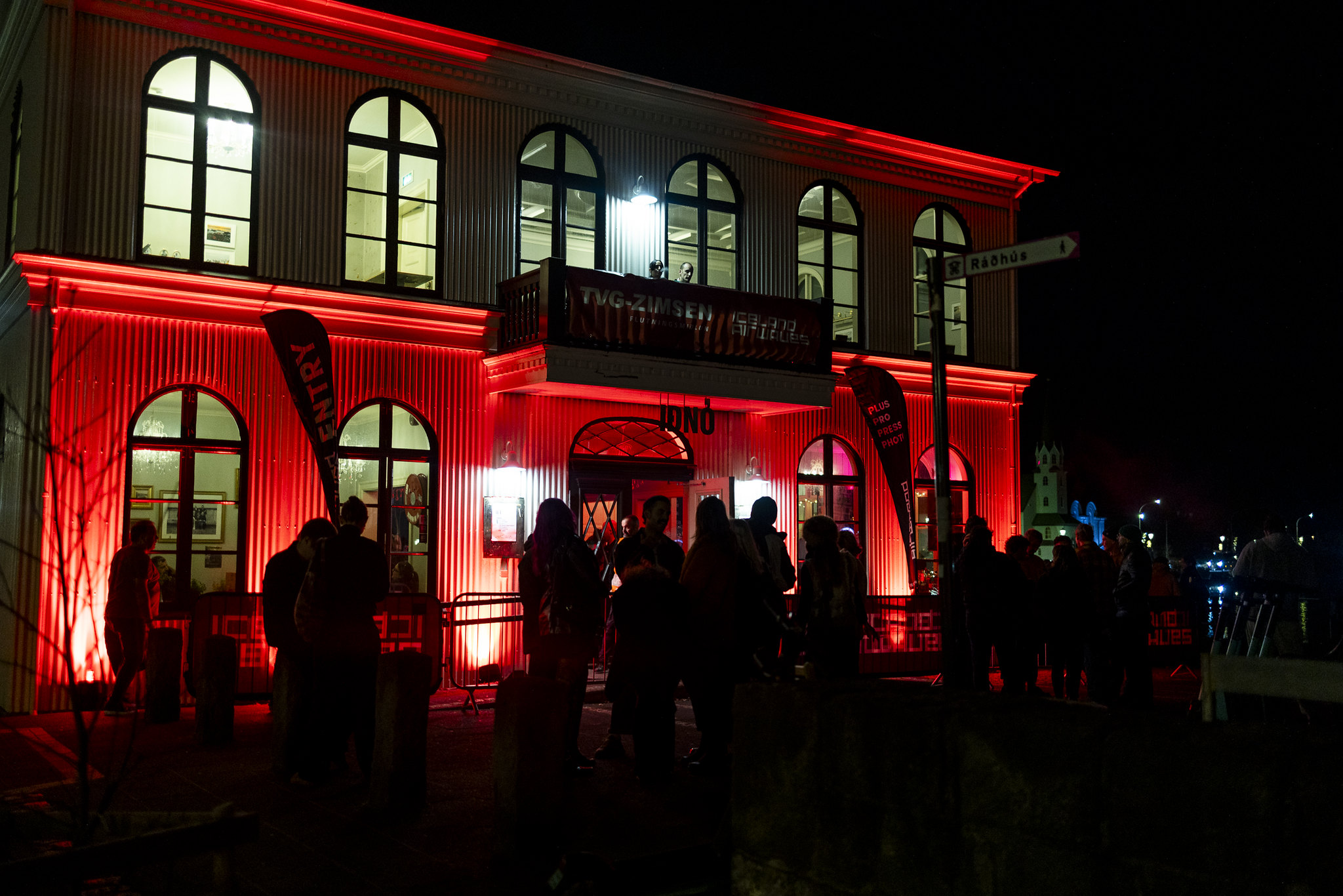 Building illuminated with red lighting at night, people gathered outside, arched windows visible on two storeys with balcony.