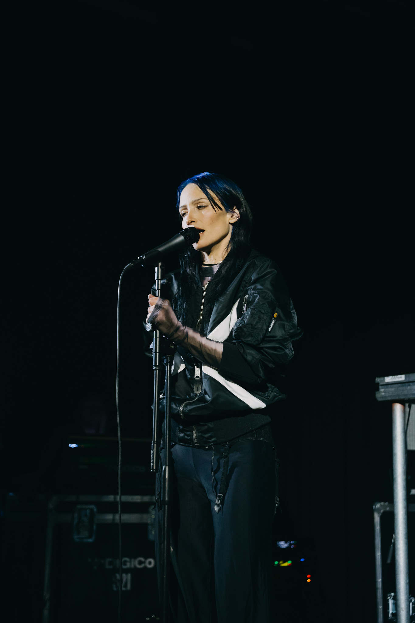 Person in black jacket and beanie singing into microphone on stage against dark background.