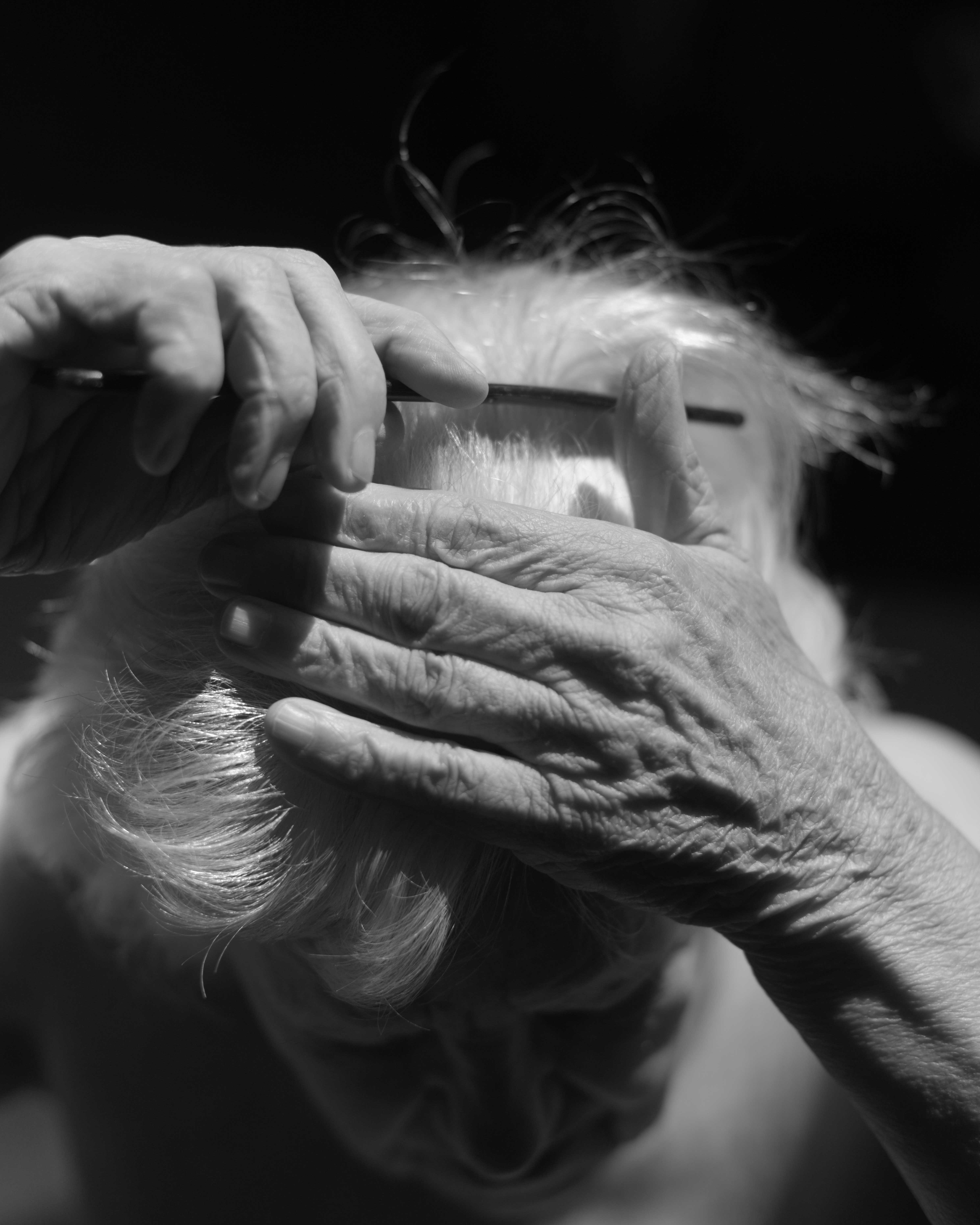 Black and white image of elderly hands using a comb on grey hair, with fine hair strands visible in the air against dark background.